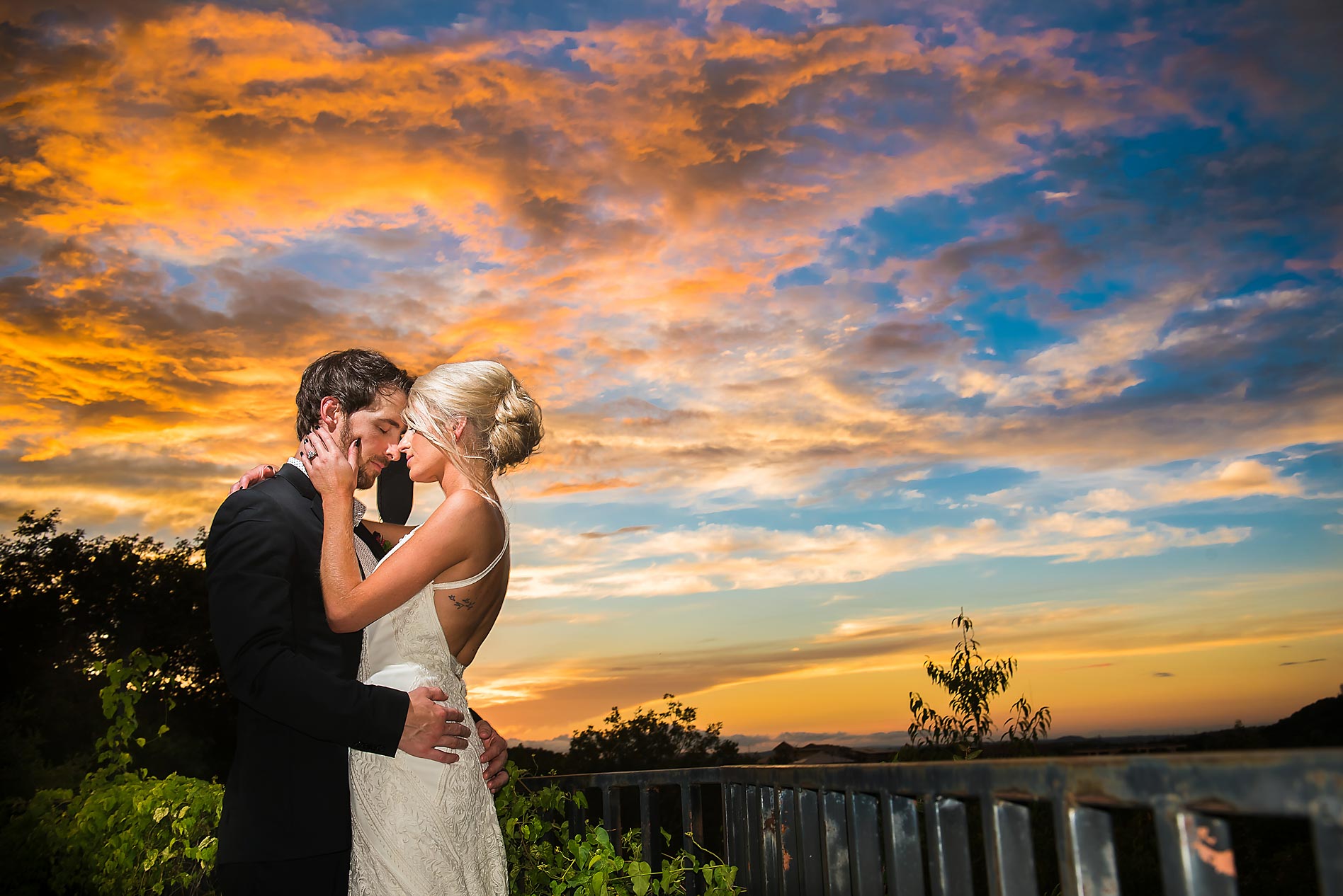 A couple in wedding attire stands close together at sunset, embracing and touching foreheads, with a colorful sky and landscape in the background.
