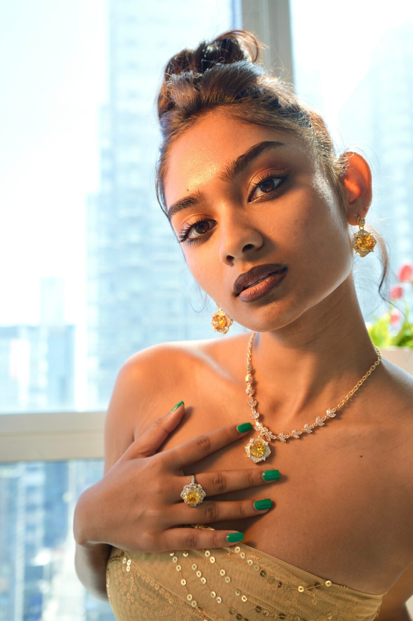 Young woman with styled hair, gold jewelry, and green nail polish, posing indoors with city buildings in background.