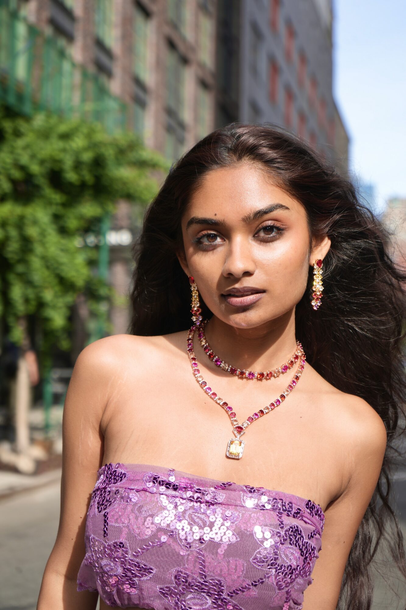 Young woman with long dark hair wearing purple sequined strapless top and jewelry, standing outdoors in city street.