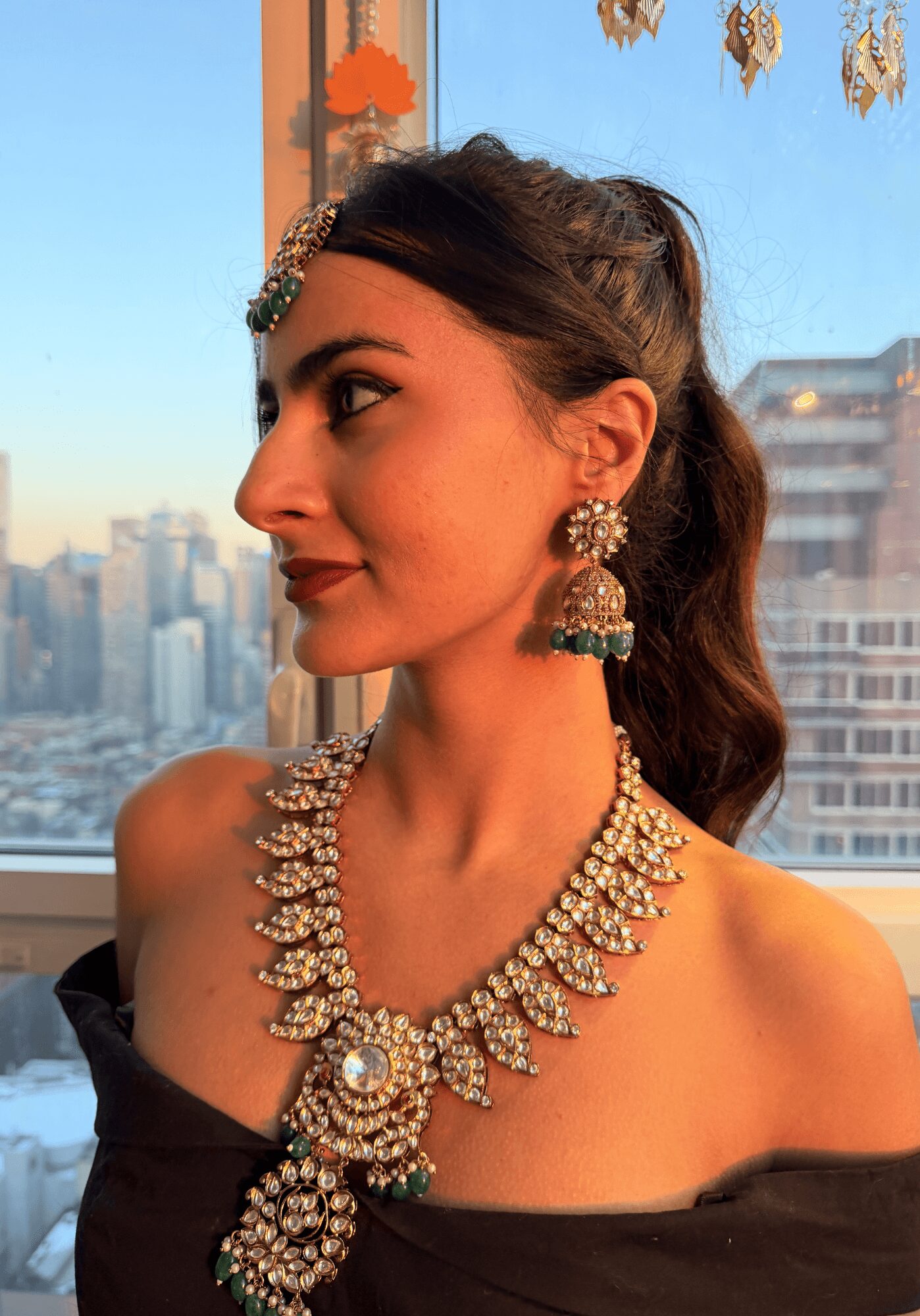 Profile of a woman wearing traditional jewelry, with cityscape background through a window.