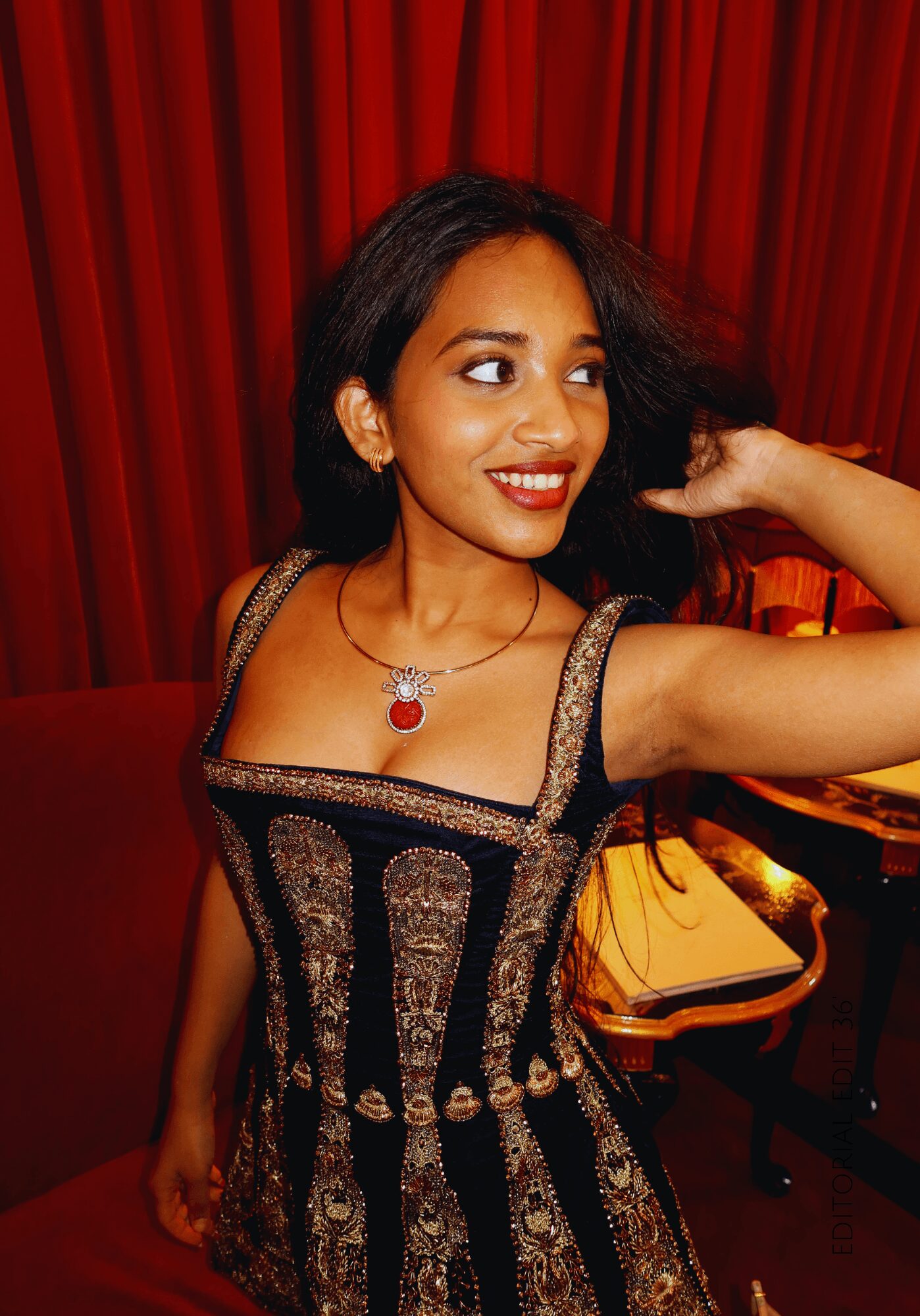 Young woman with dark hair smiling, wearing a black and gold dress, in front of red curtains.