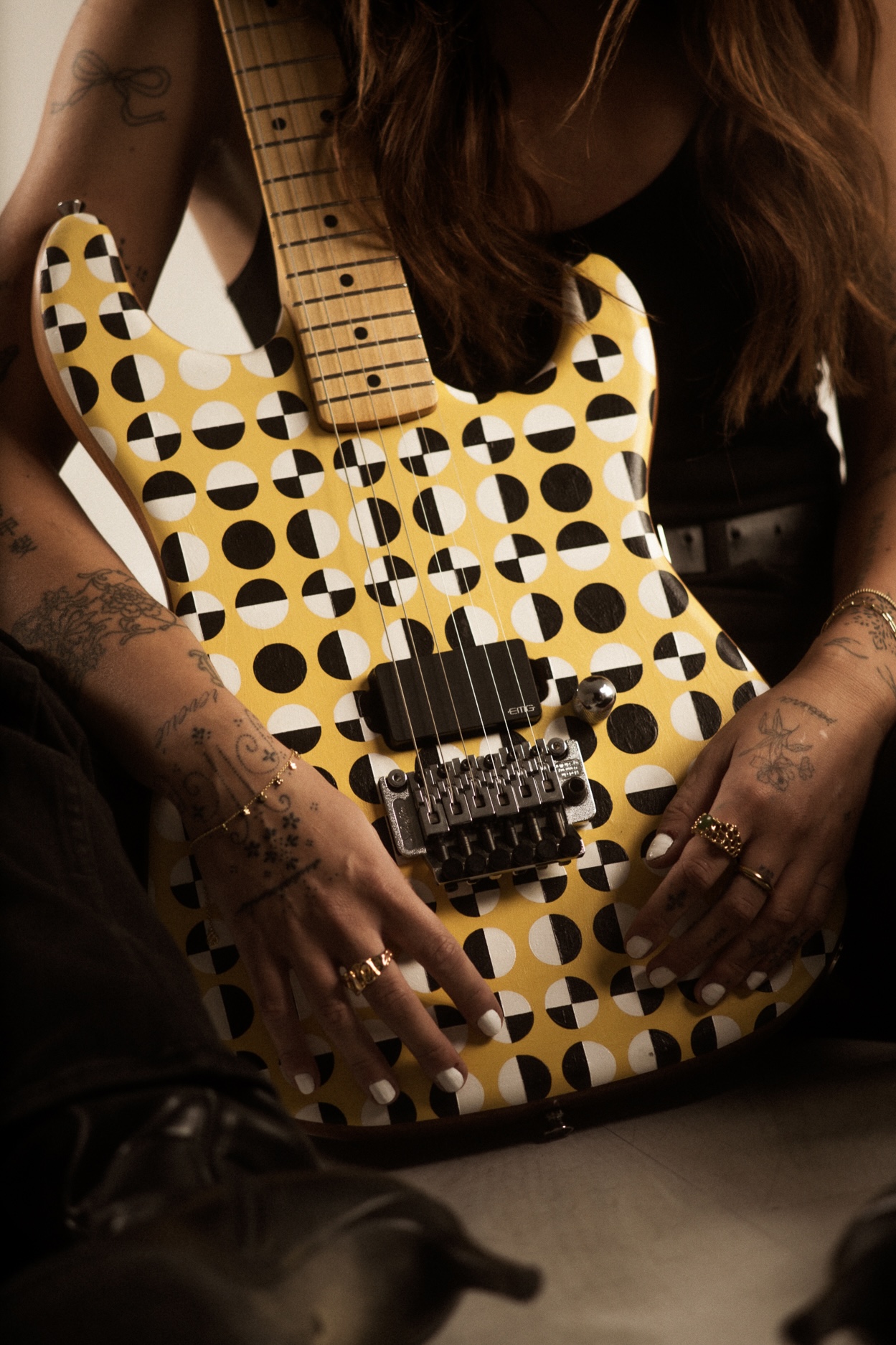 Person holding a yellow and black patterned electric guitar with a wooden neck, tattoos on arms, rings on fingers.