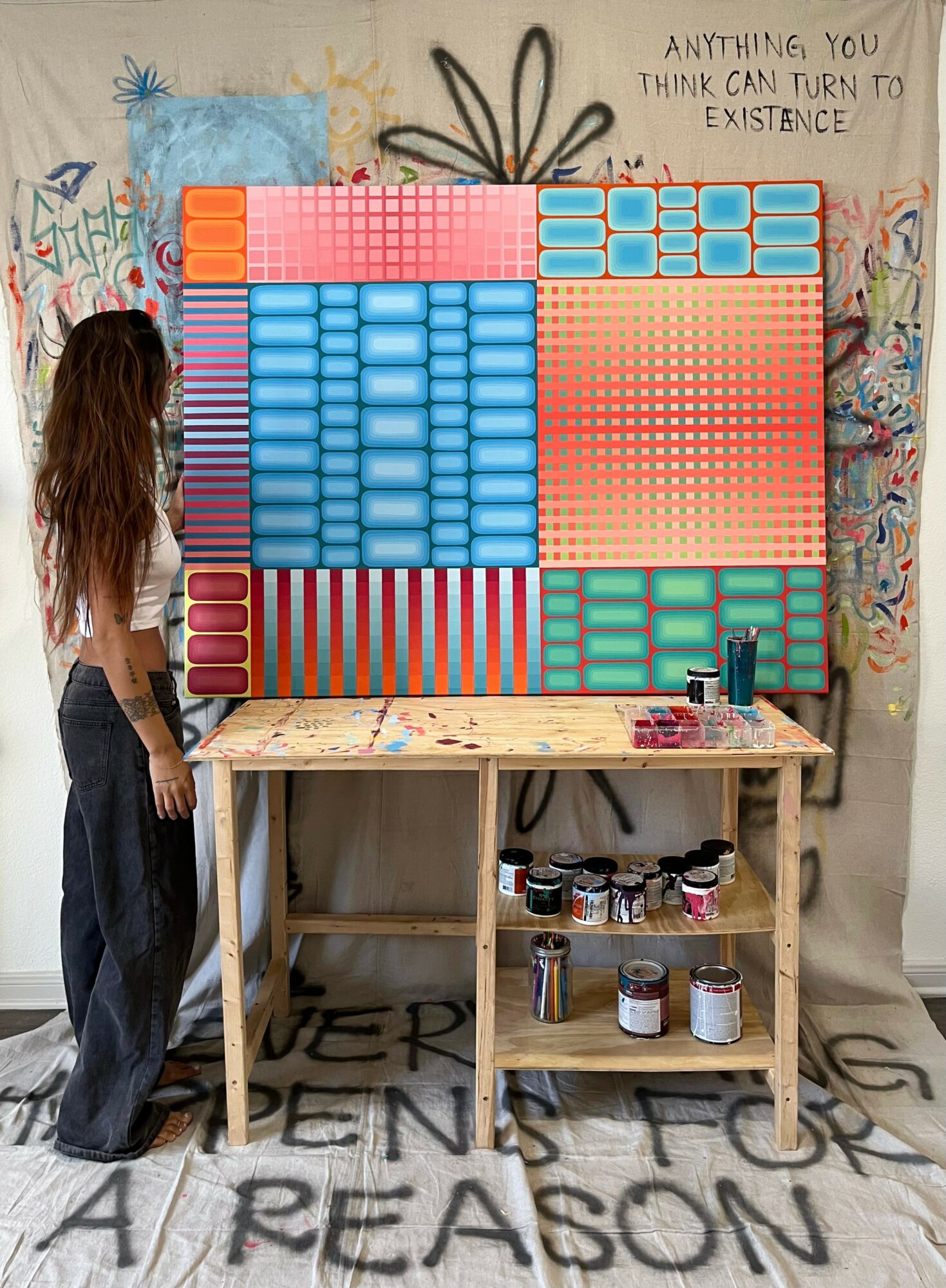 Woman with long hair standing in front of a colorful pixel art display on a wall, with a wooden table and art supplies below. Word graffiti on wall and floor.