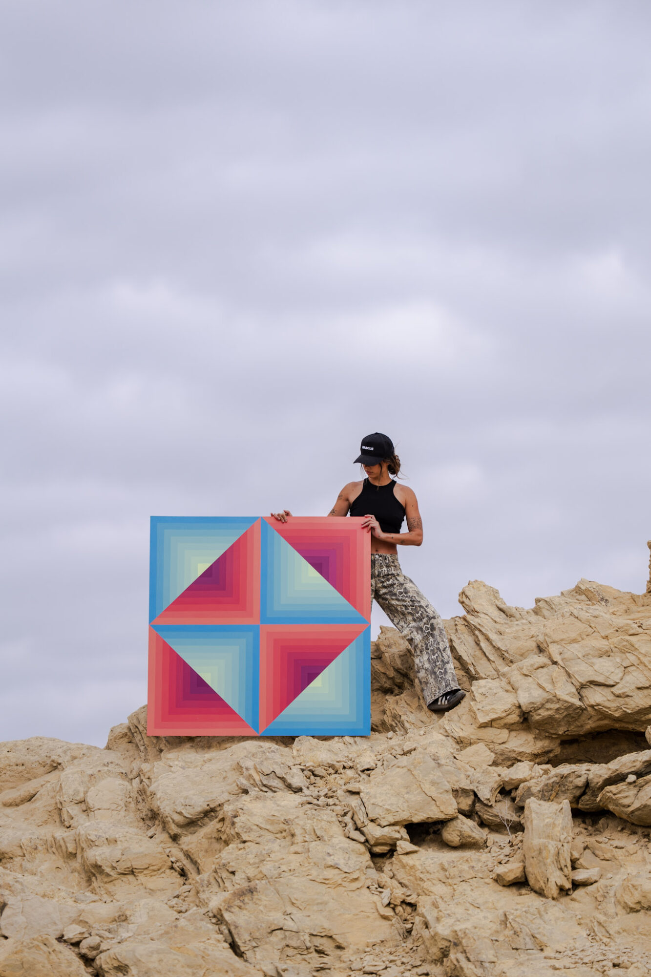 Person sitting on rocky terrain with a colorful geometric sculpture, cloudy sky in background.