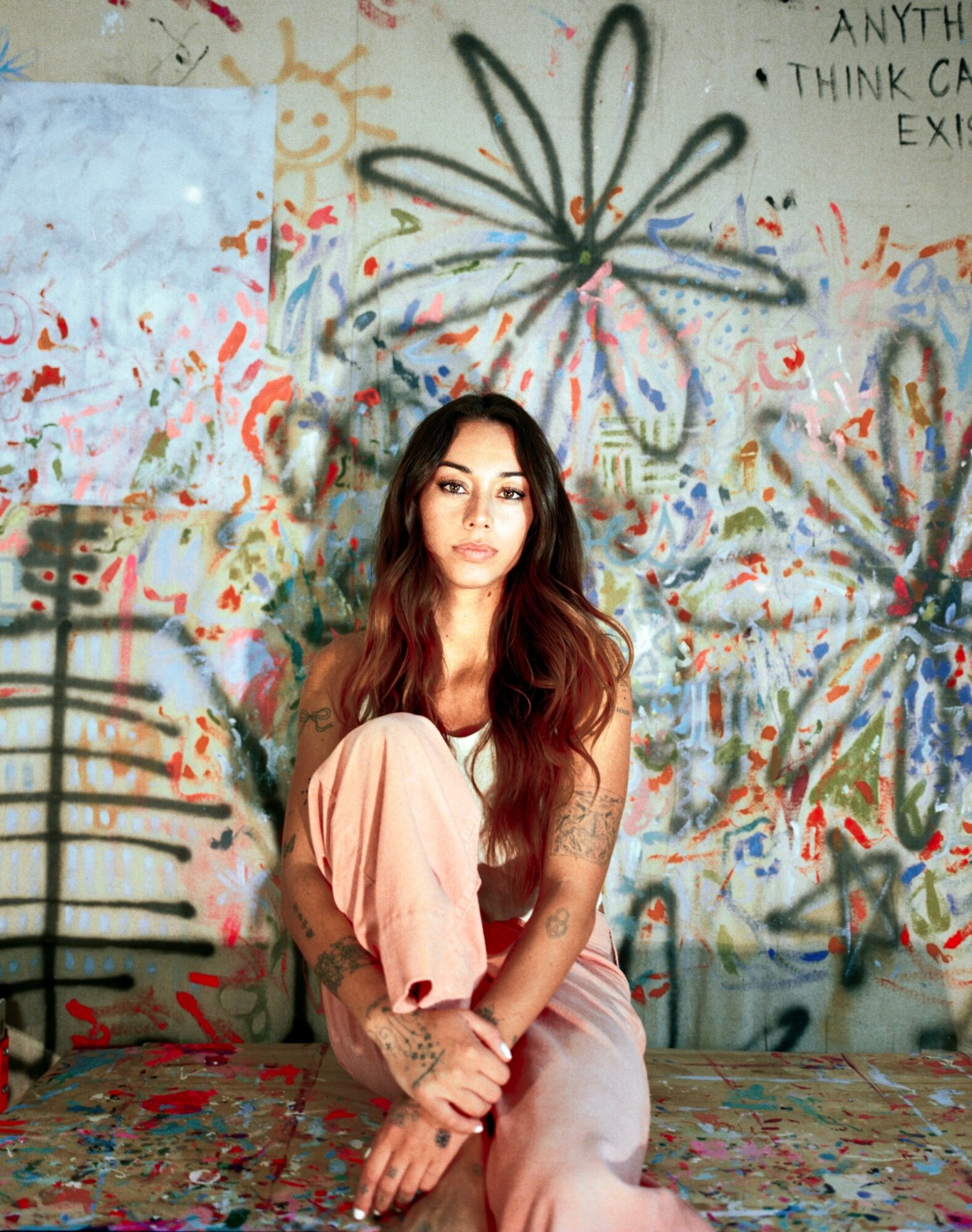 Young woman with long hair sitting on a colorful, graffiti-covered wall background, wearing a light-colored outfit, looking at the camera.