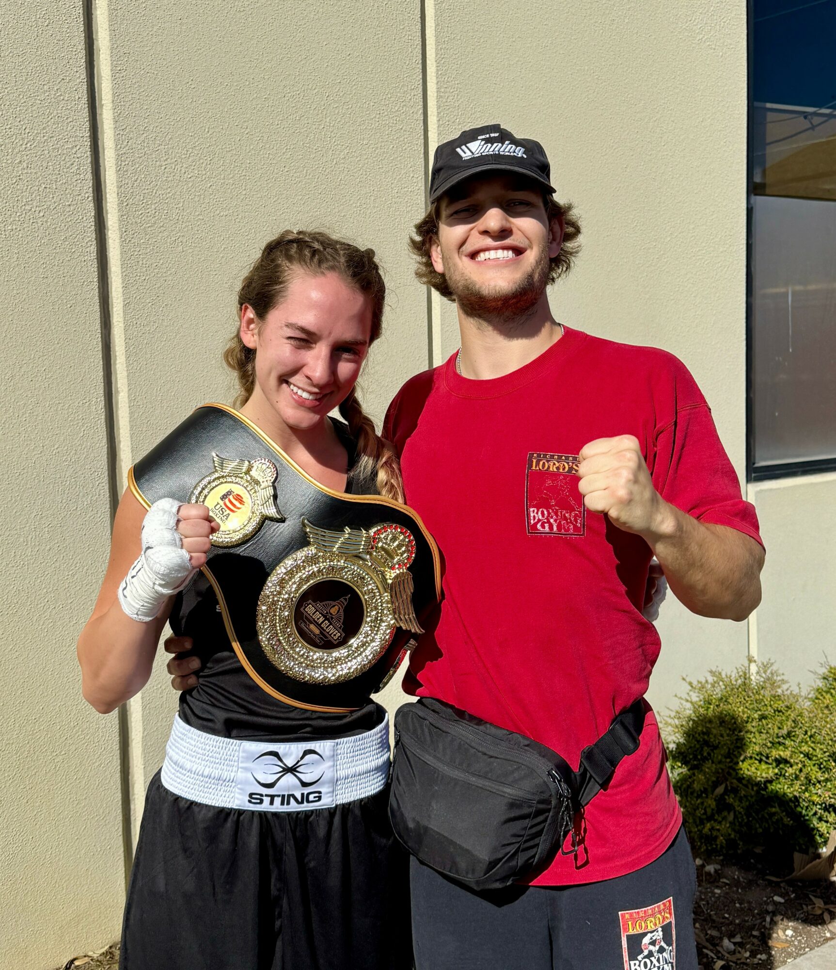 Two young people smiling, one holding a championship belt, outdoors near a building, sunny day.