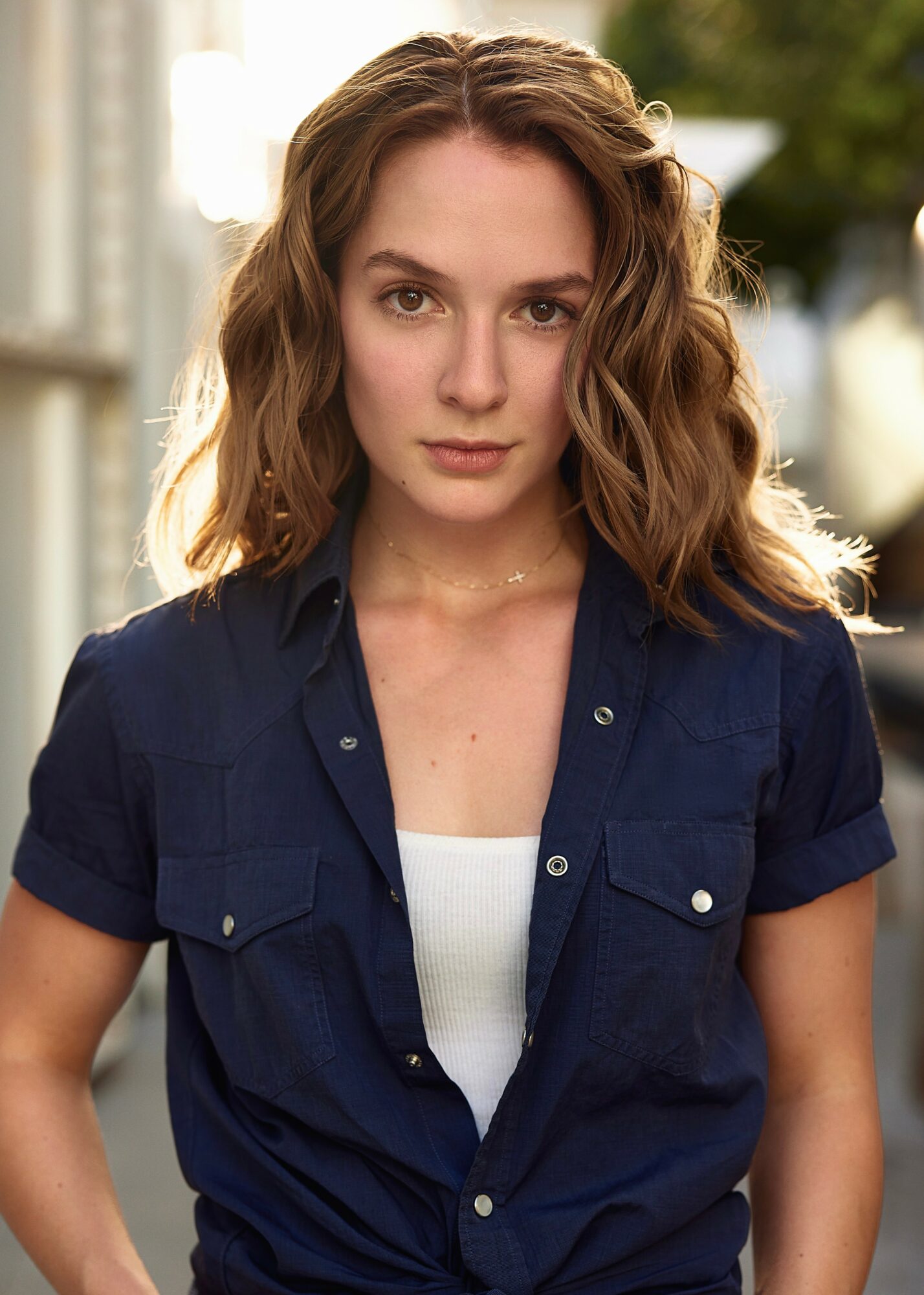 Young woman with wavy brown hair wearing a navy shirt outdoors, looking at the camera.