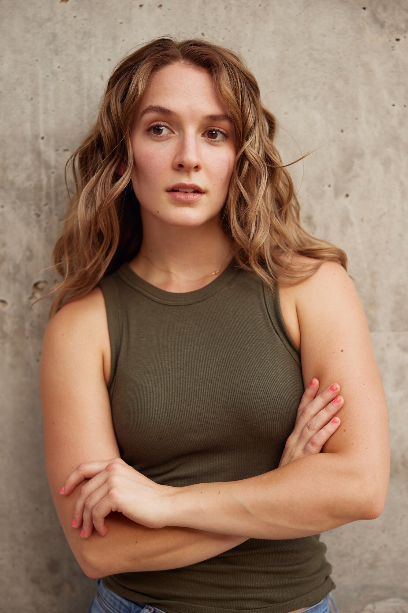 Young woman with wavy hair wearing a sleeveless olive green top, standing against a concrete wall, arms crossed.