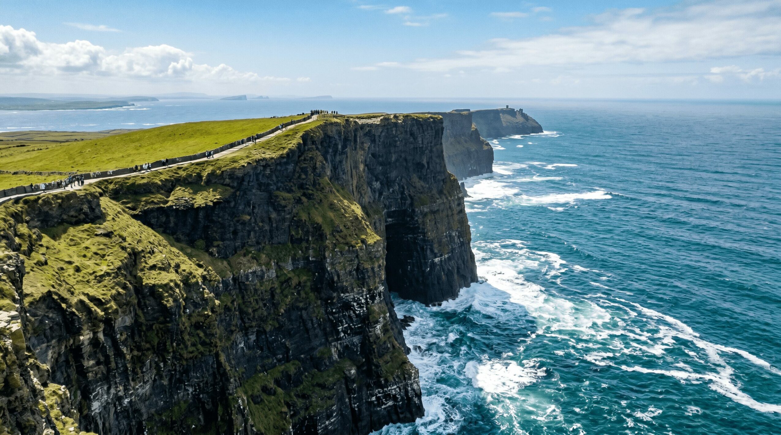 Cliffs along a coastline with green grass and ocean waves, under a partly cloudy sky.