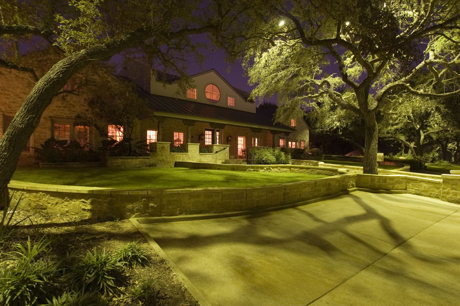 Night view of a house with lit windows, surrounded by trees and a curved pathway, illuminated by outdoor lighting.