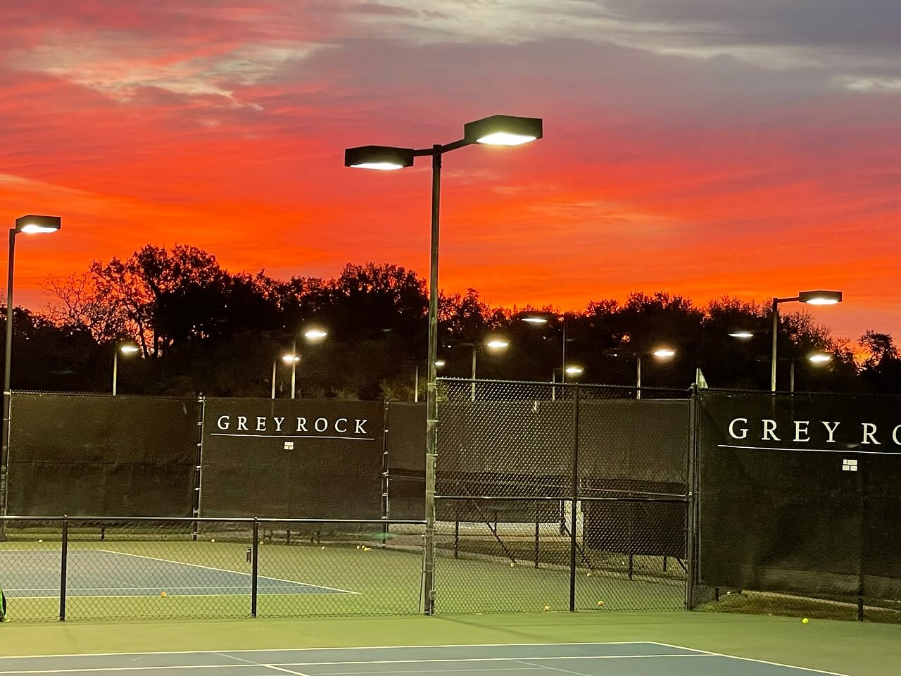 Tennis courts with black wind screens, illuminated by tall lights, against a vibrant sunset sky with orange and pink hues.