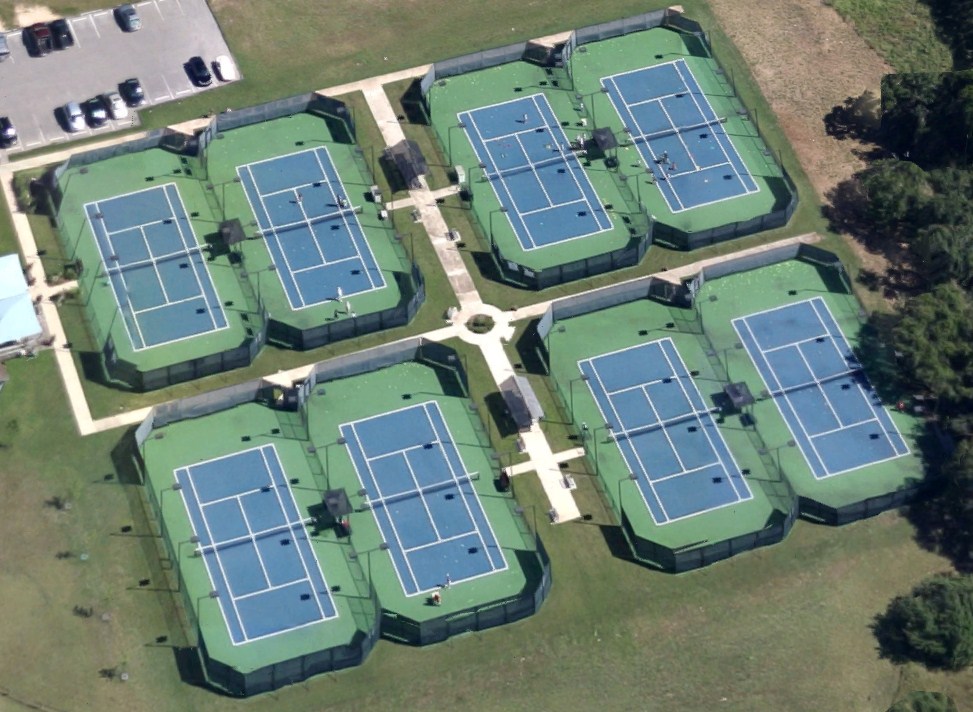 Eight tennis courts with surrounding green fences arranged in two rows, connected by pathways, with parking lot nearby.