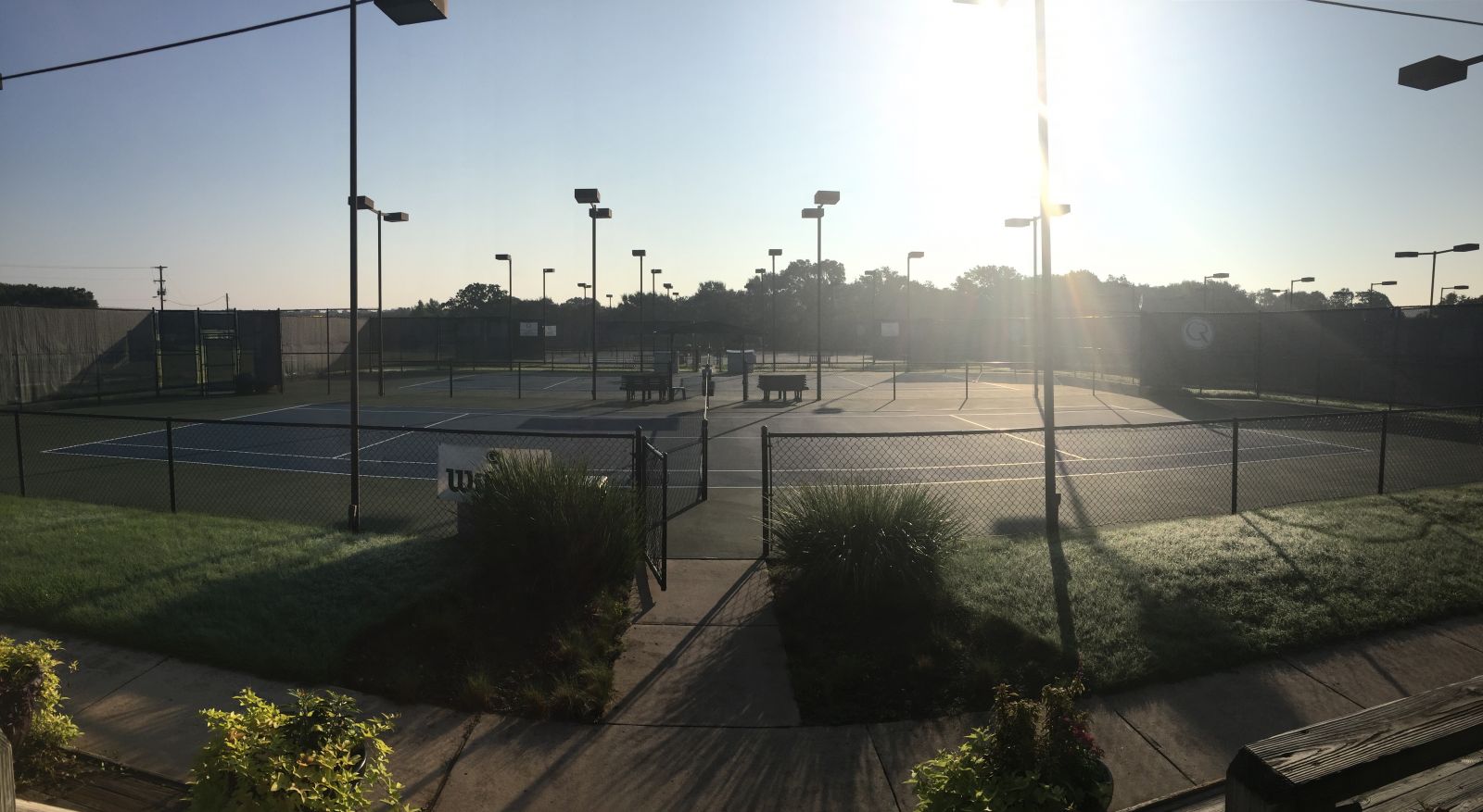 Empty outdoor tennis courts with surrounding fencing, light poles, and a pathway, under bright sunlight.