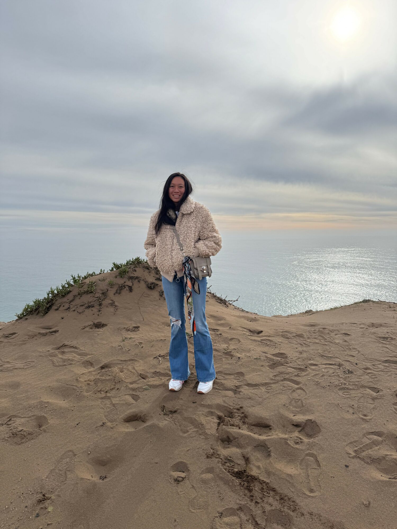 Person standing on sandy beach near water, wearing a beige jacket and blue jeans, with footprints in the sand.