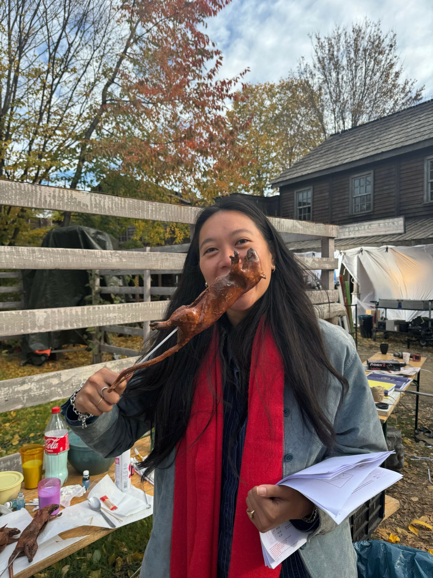Person with long dark hair wearing a gray coat and red scarf, holding a large cooked meat skewer, outdoors with trees and building in background.