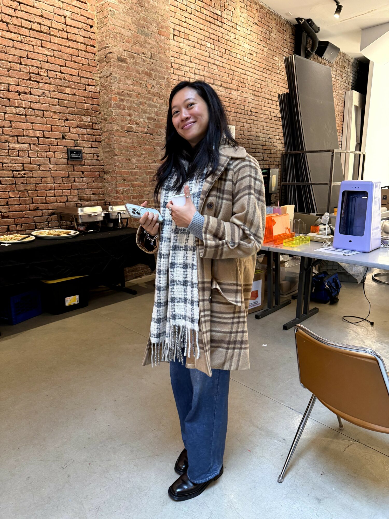 Woman standing indoors, smiling, wearing a plaid coat and scarf, with a brick wall background.