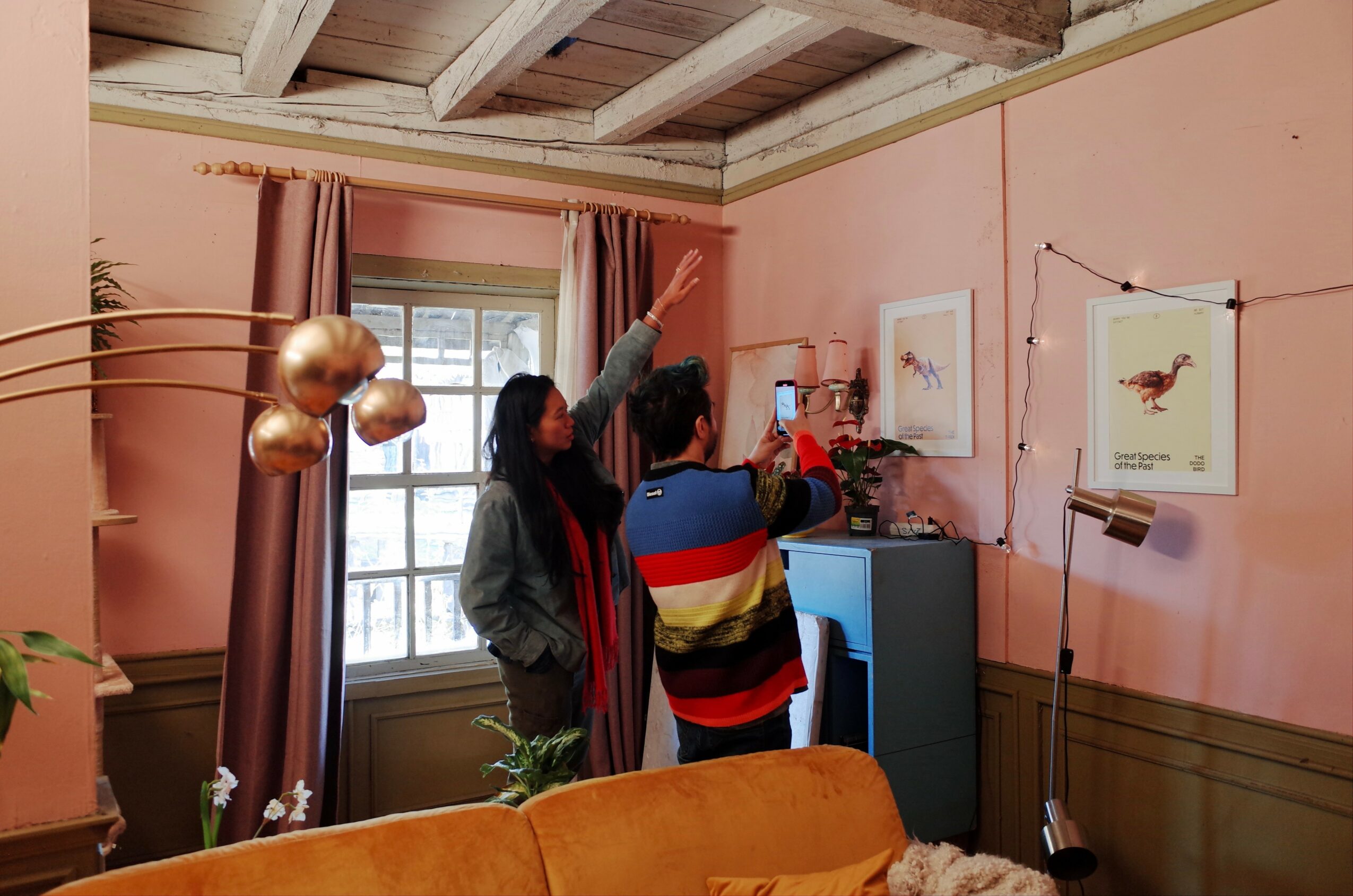 Two people stand in a pink-walled room with artwork, a window, and a blue cabinet, taking a photo together.