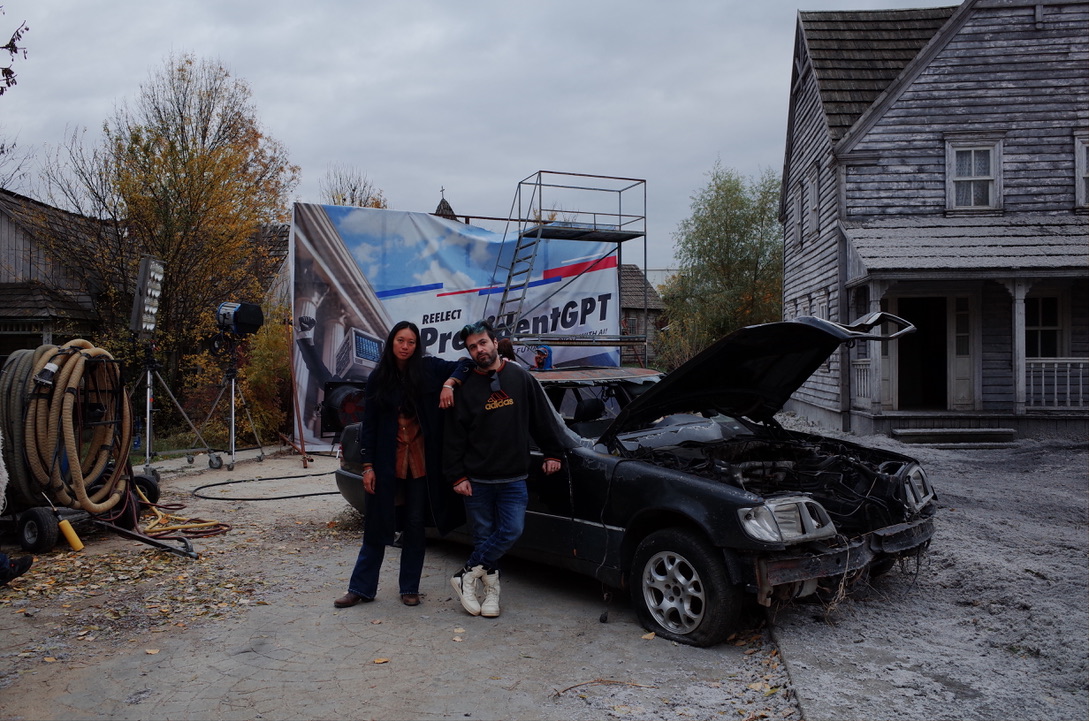 Two people stand next to a damaged black car with an open hood, in front of a building and a billboard, on a cloudy day.