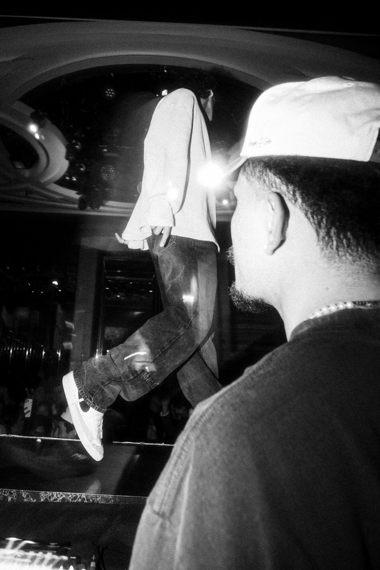 Person wearing a cap and glasses looking at a person walking inside a vehicle at night, with bright light reflections.