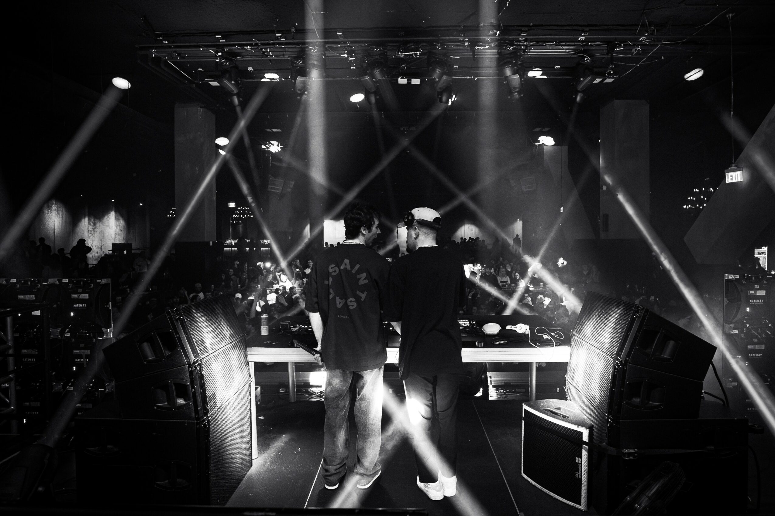 Two people stand at a DJ booth in a dark, illuminated nightclub with bright stage lights and a city skyline backdrop.