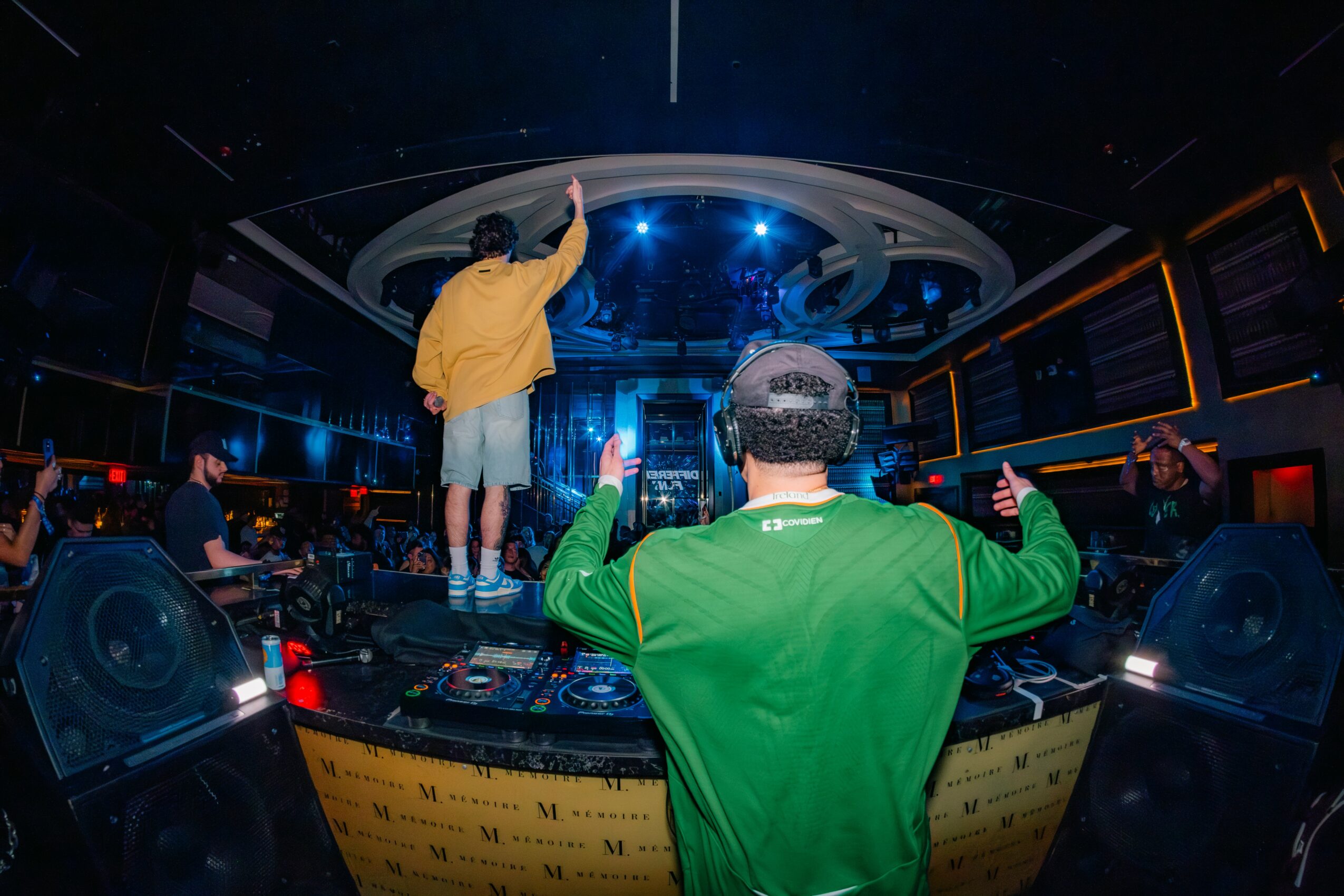 Person in green jacket with headphones facing DJ booth, another person on stage reaching towards ceiling, nightclub setting with lights.