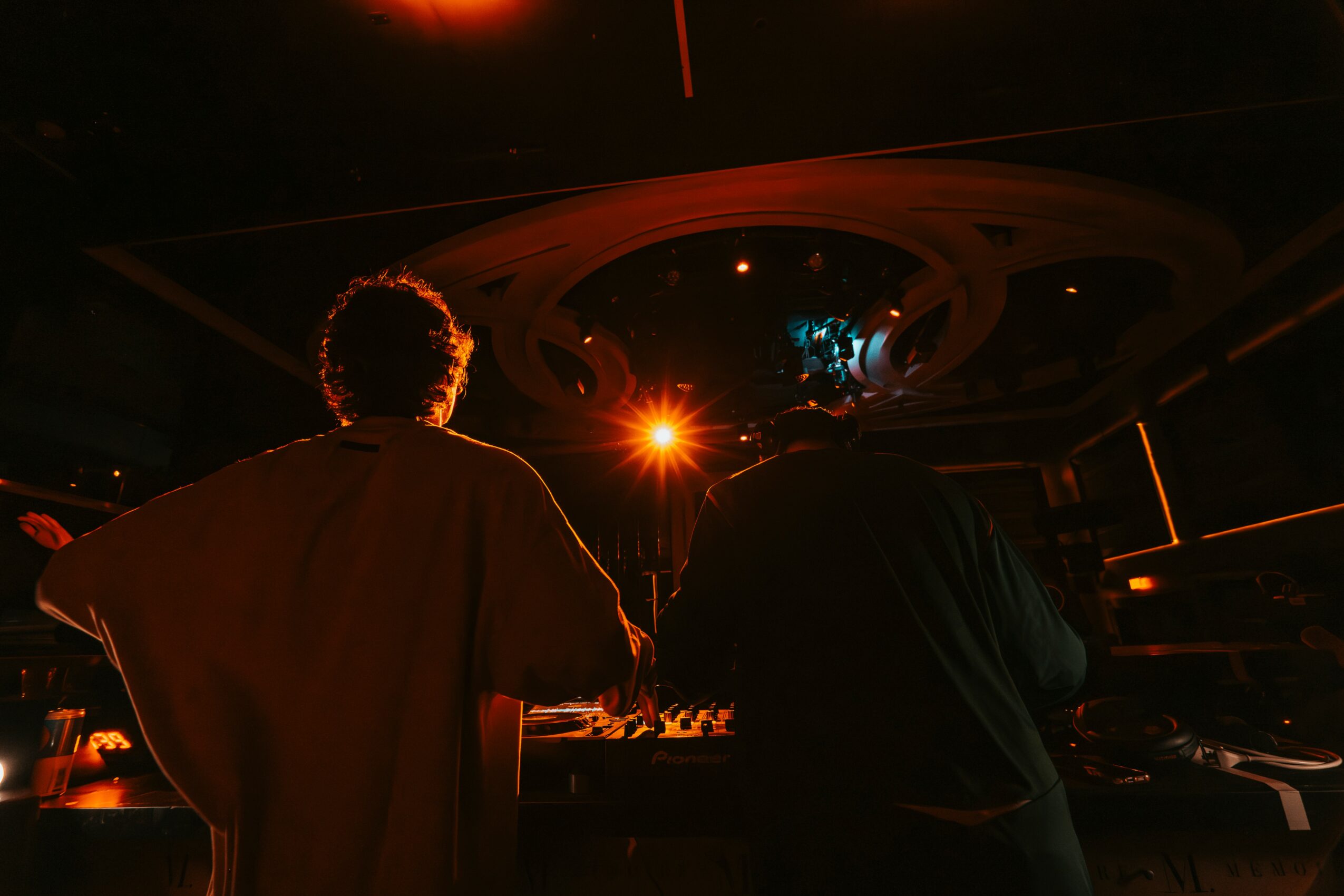 Two people are seen from behind in a dark room with a ceiling light fixture, illuminated by a bright orange light.