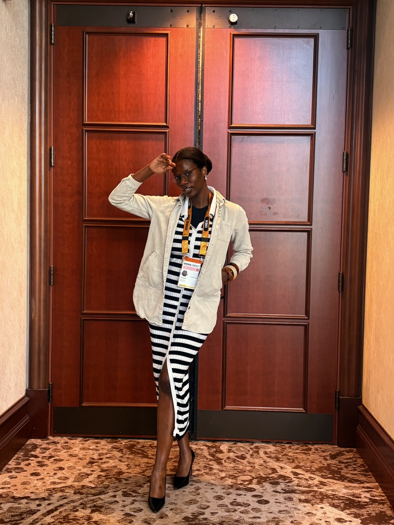 Woman standing in front of wooden doors, wearing a striped dress, beige coat, and high heels, holding a camera, indoors.