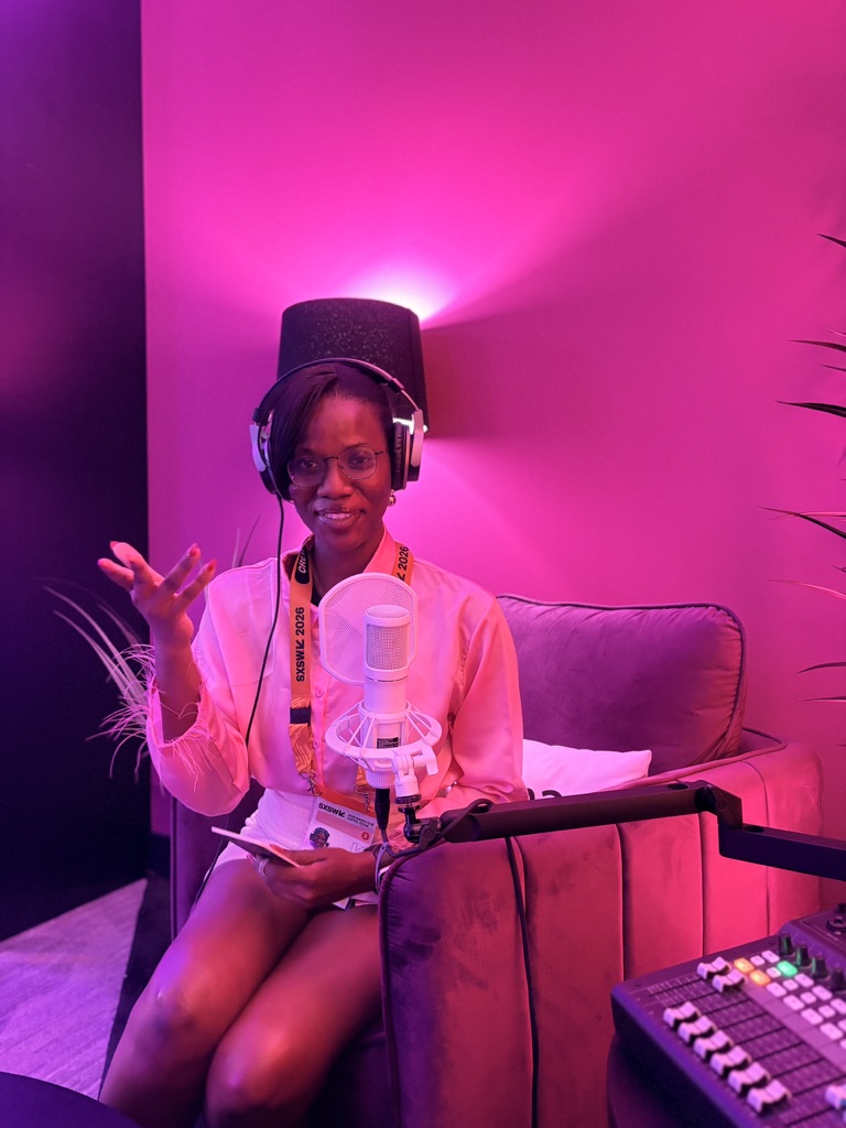 Young woman in pink shirt with headphones, sitting on a purple couch in a recording studio with pink lighting, holding a notebook.