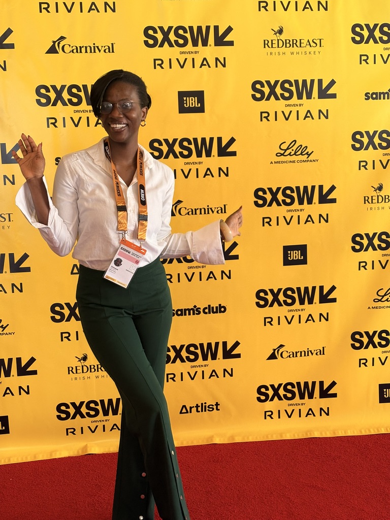 Woman smiling and waving at SXSW event with yellow backdrop, wearing glasses, a beige blazer, and dark pants.