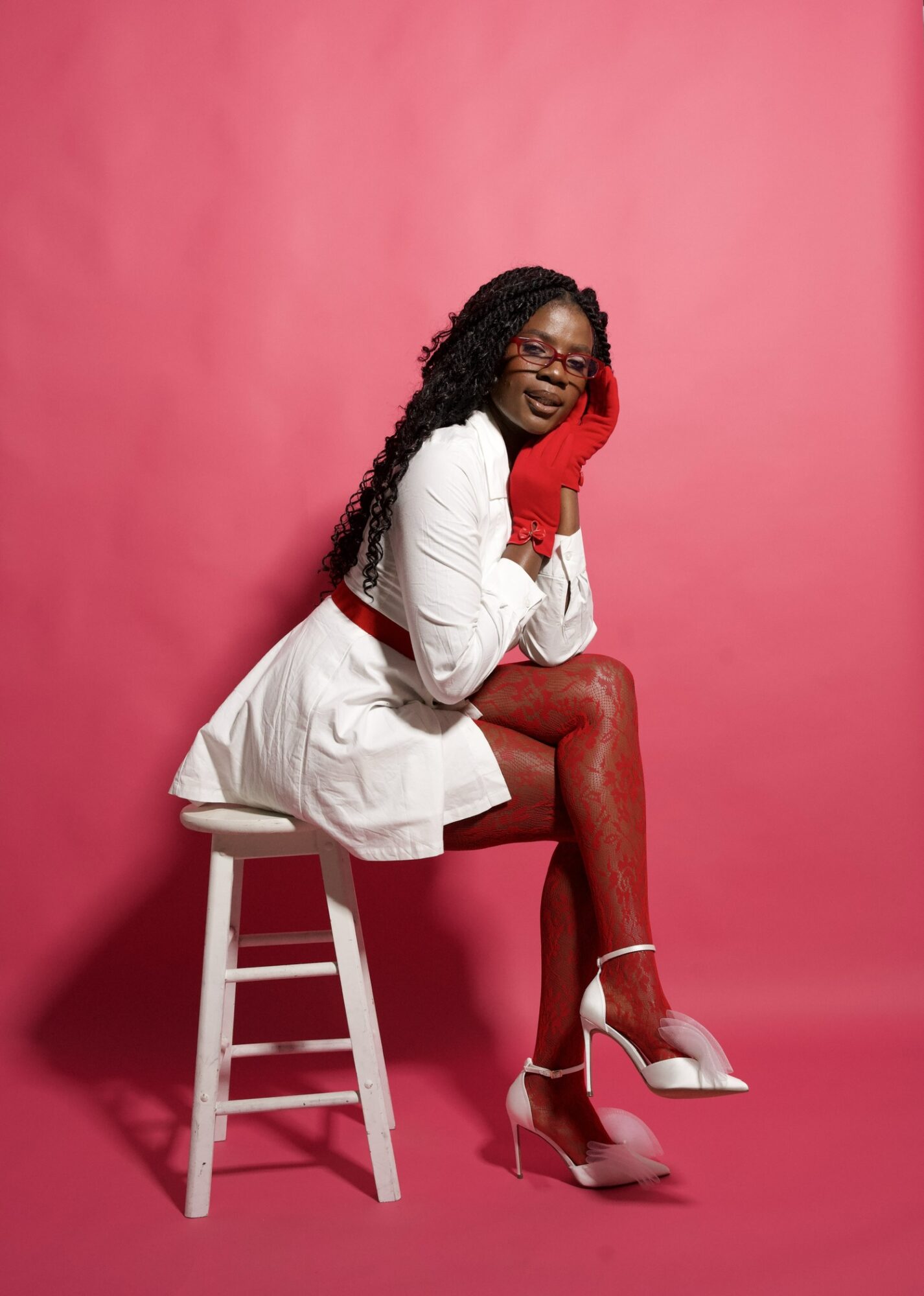 Woman with long curly hair, glasses, white dress, red gloves, red tights, white heels, sitting on a stool against pink background.