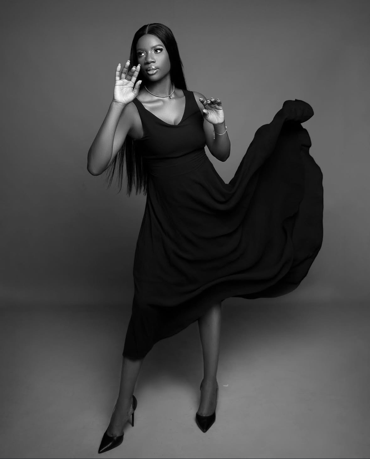 Woman in a long dress with one arm raised, standing against a plain background.