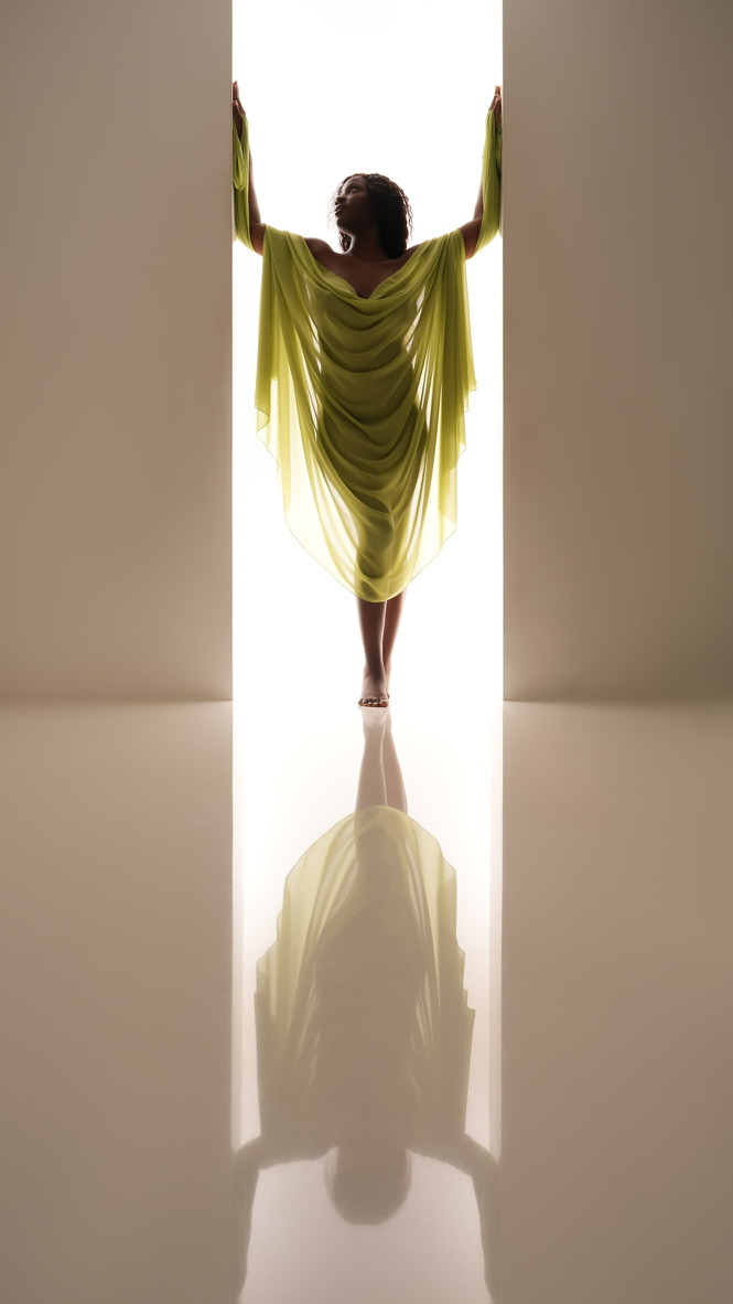 Person standing in a doorway with arms raised, wearing a flowing yellow dress, with reflection on the floor.