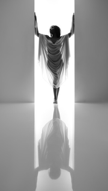 Person standing in a doorway with arms raised, wearing a flowing dress, reflected on the floor, black and white image.