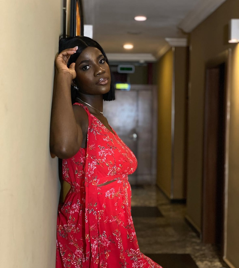Woman in red dress leaning against wall in hallway, looking at camera, with hallway background.