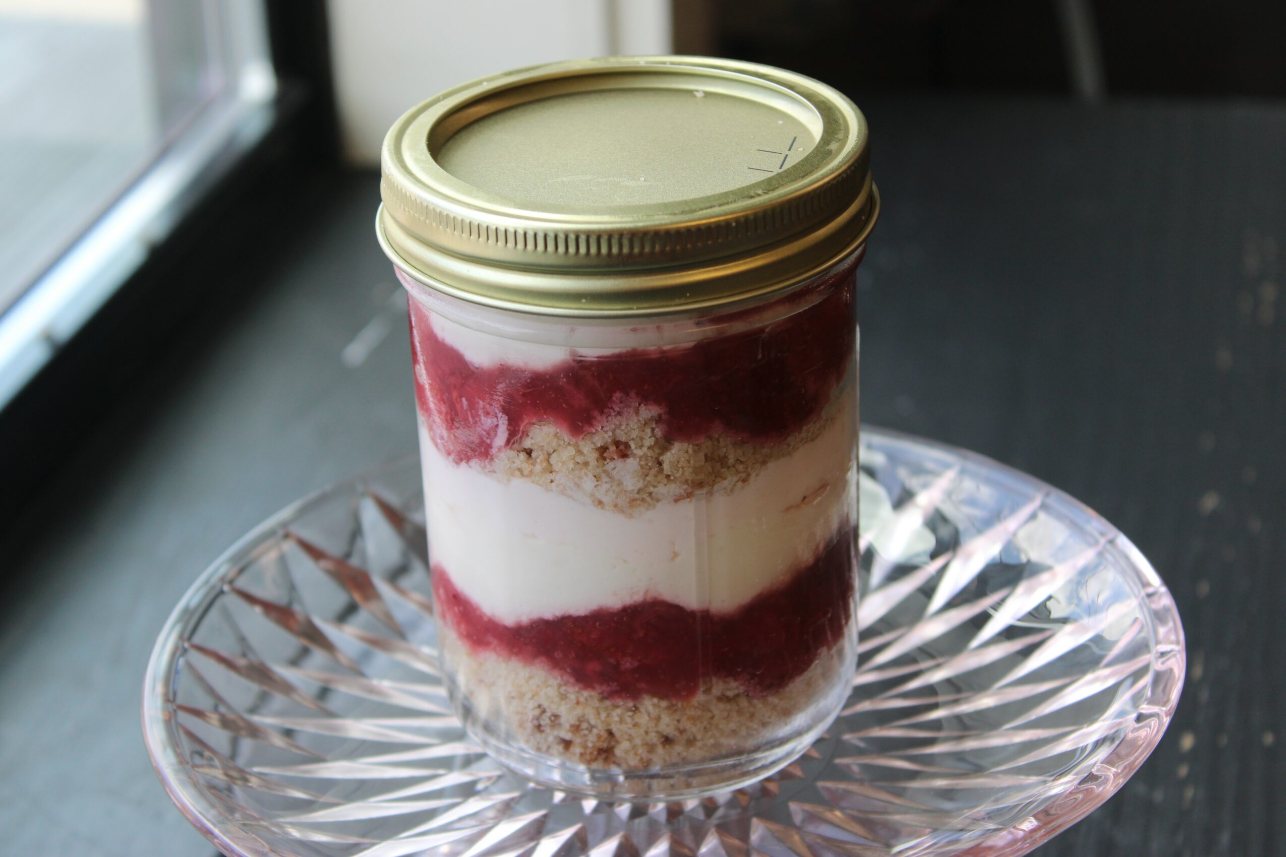 Layered dessert in a jar with cream, red fruit sauce, and cake, on a glass plate near a window.