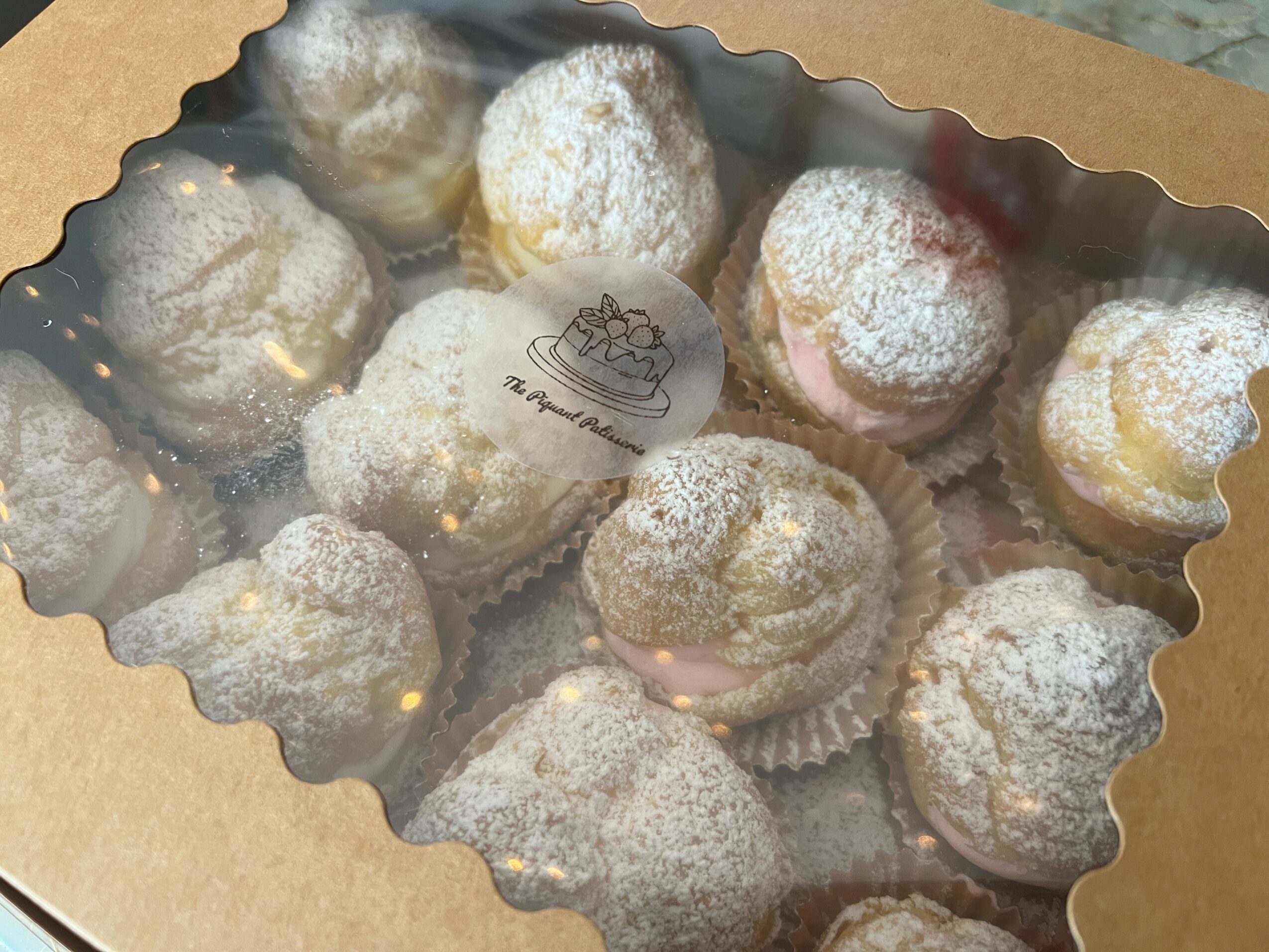 Box of powdered sugar-coated cream puffs or profiteroles with a small label inside, viewed through a clear plastic window.