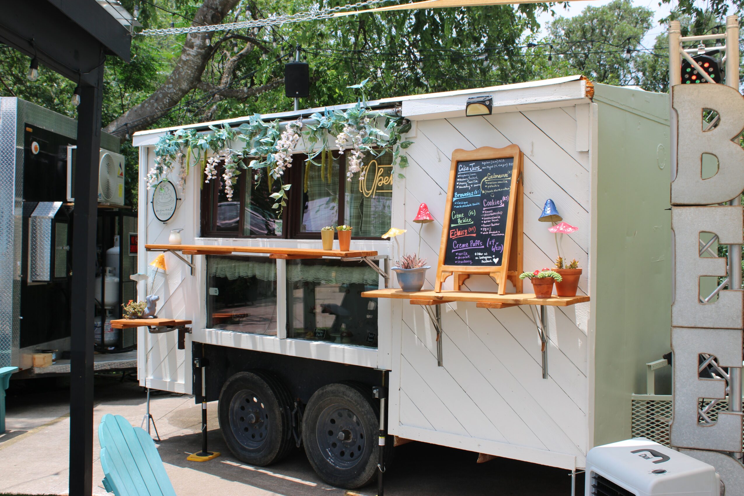 Food truck with a chalkboard menu, decorative plants, and outdoor seating, surrounded by trees and other structures.