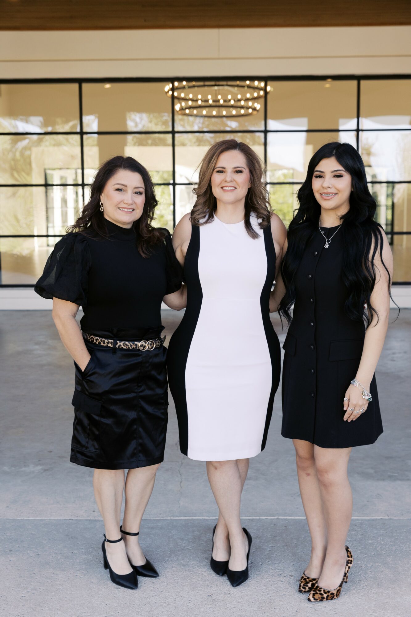 Three women standing indoors in front of large glass windows, smiling, dressed in black and white dresses, with a chandelier above.