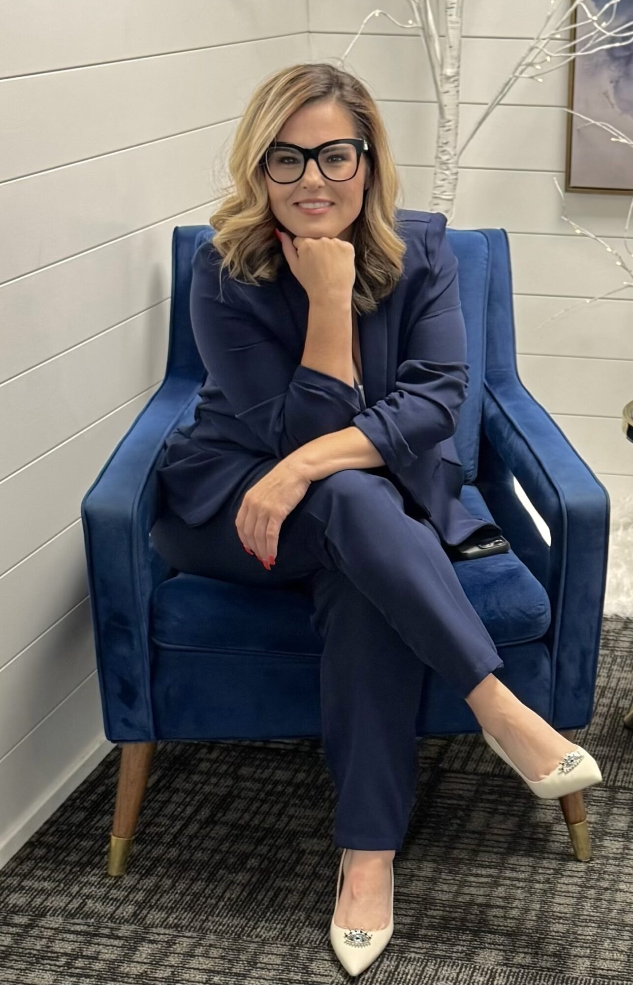 Woman with glasses sitting on a blue armchair, smiling, in a room with white walls and a patterned carpet.