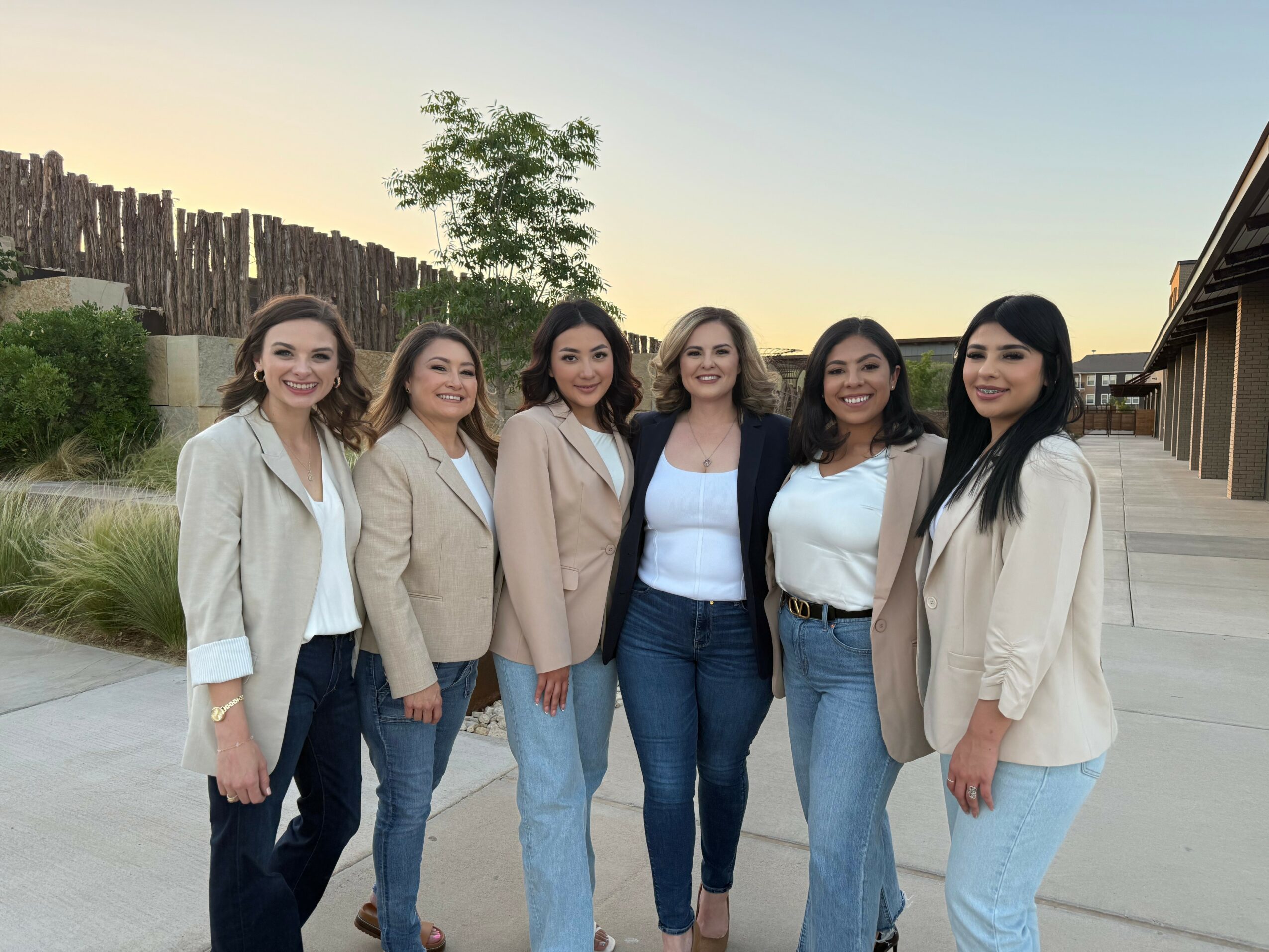 Six women standing outdoors, smiling, with trees and buildings in the background.