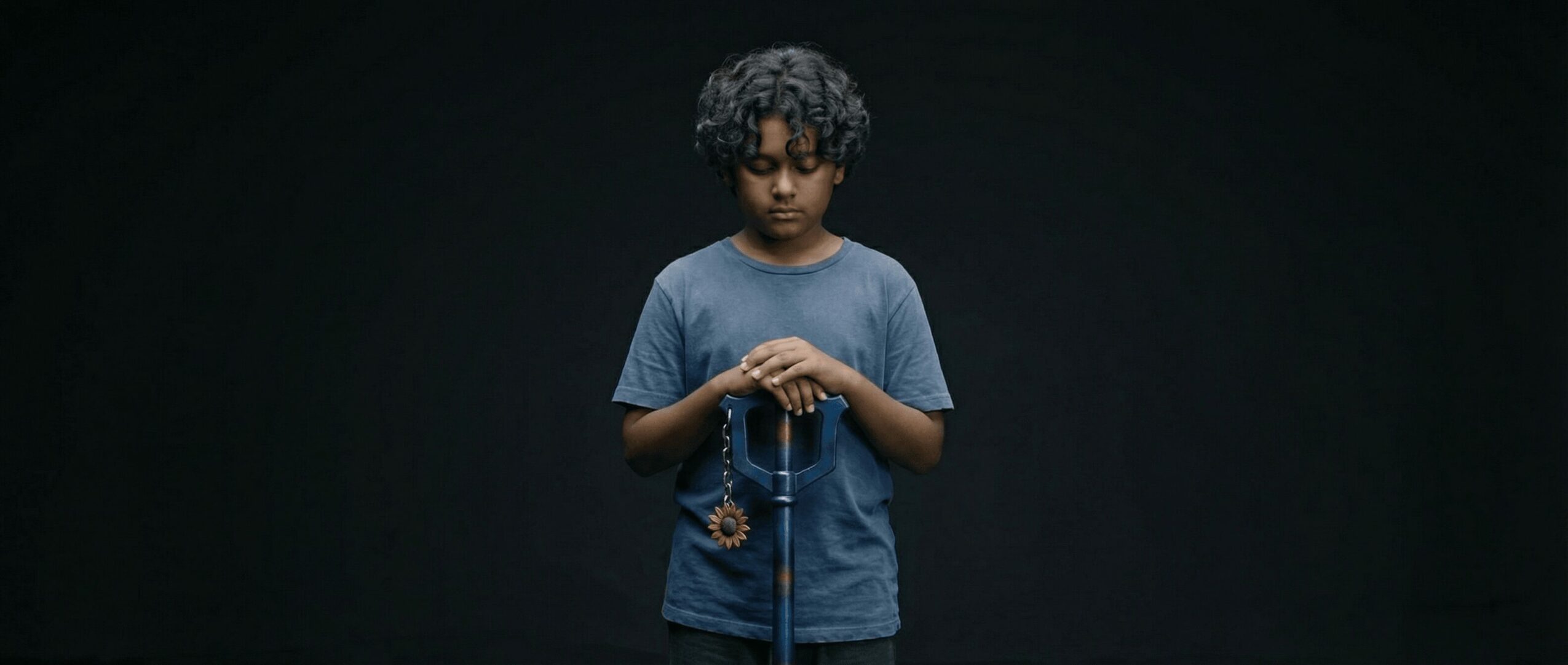 Young boy with curly hair wearing a blue t-shirt, holding a necklace, standing against a dark background.