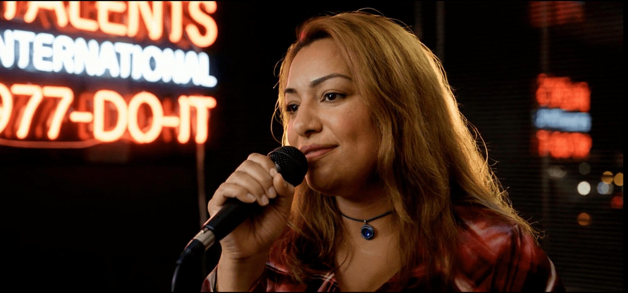 Woman singing into microphone on stage with neon signs in background, smiling, with long hair and a necklace.