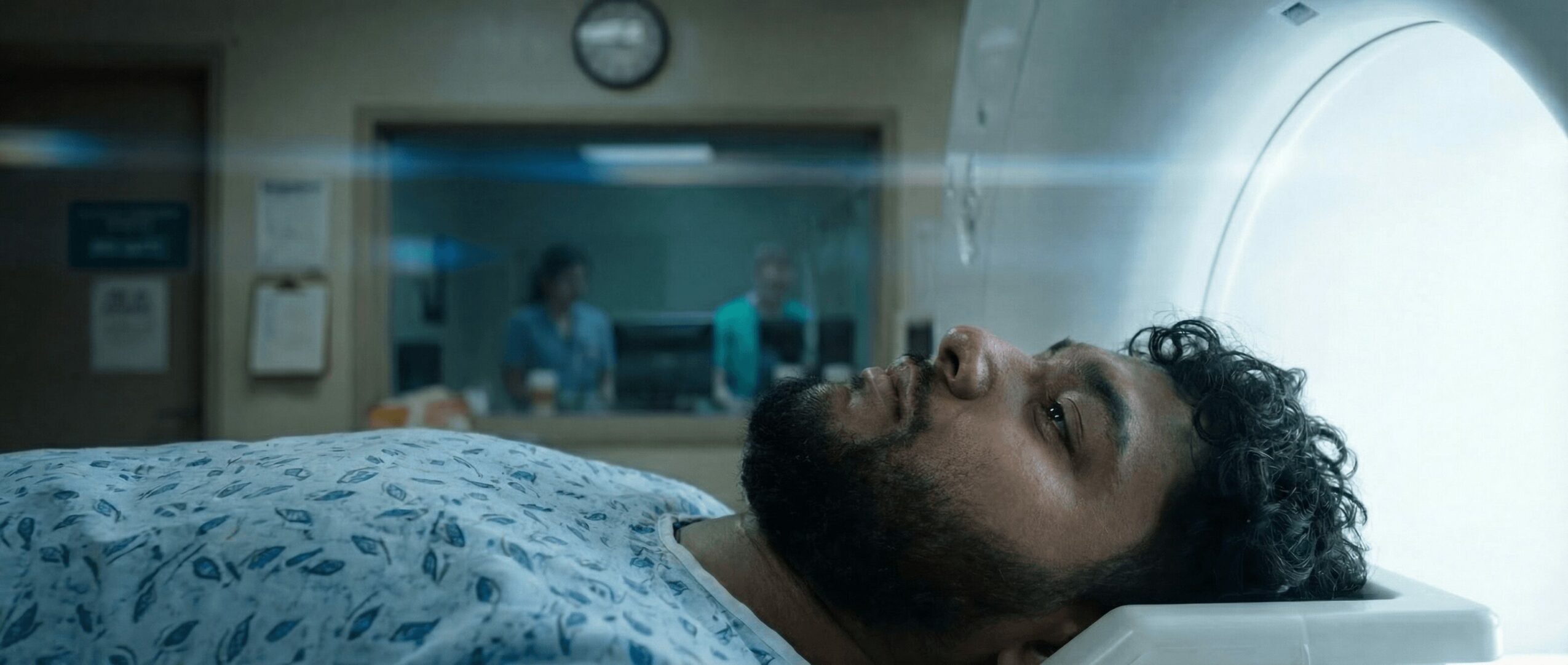 Man lying on hospital bed with medical staff visible through window in background, medical equipment nearby, patient appears to be undergoing a scan.