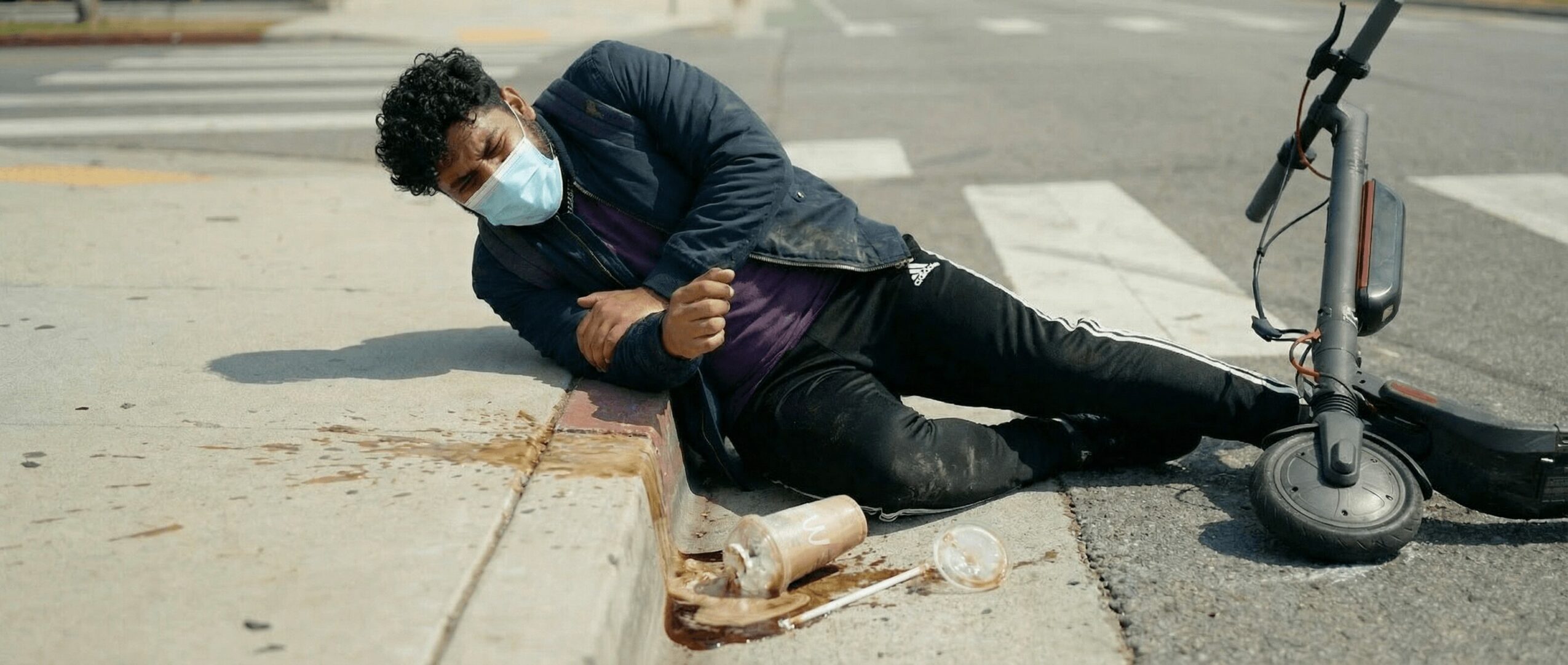 Person lying on the sidewalk, wearing a mask, next to an electric scooter, with a plastic bottle nearby.