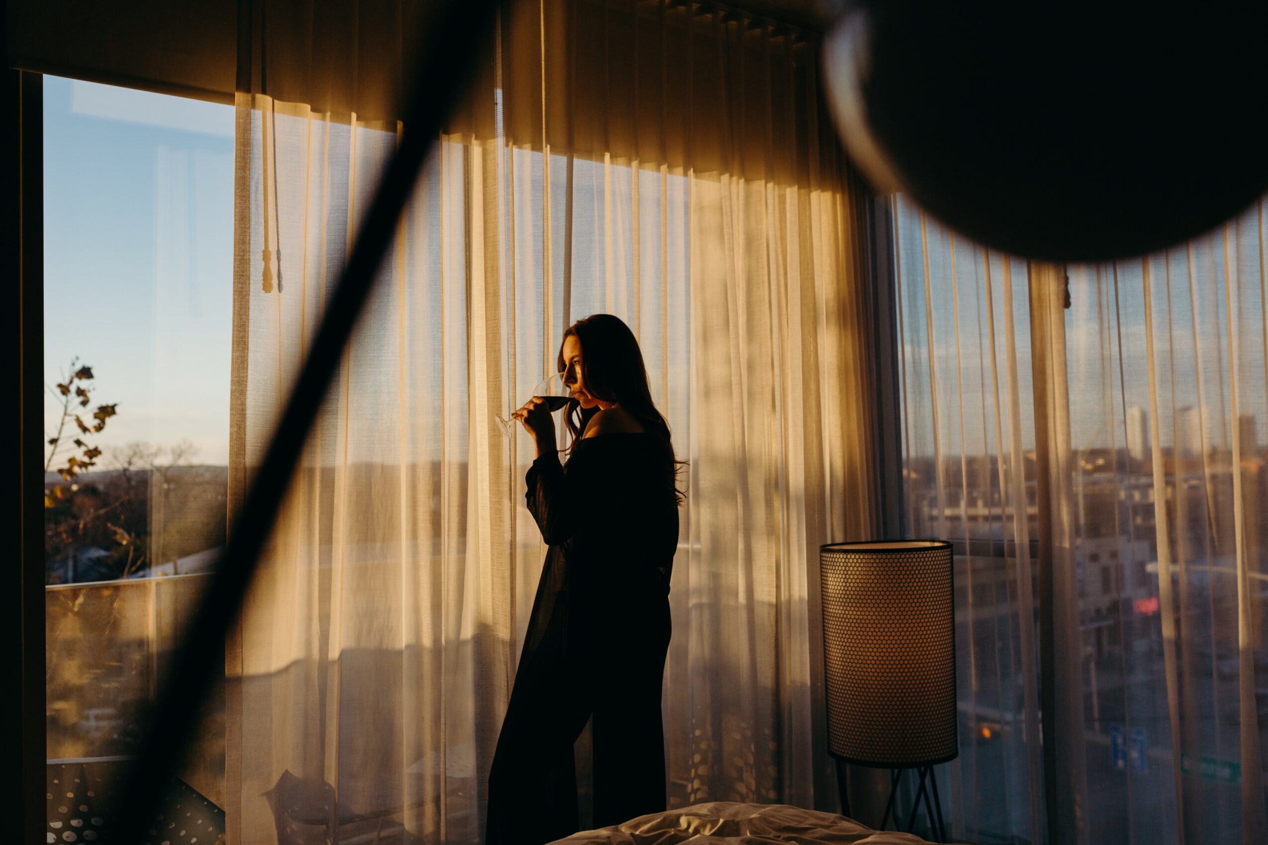 Person standing by window with curtains, holding a drink, looking outside during sunset, cityscape visible through window.