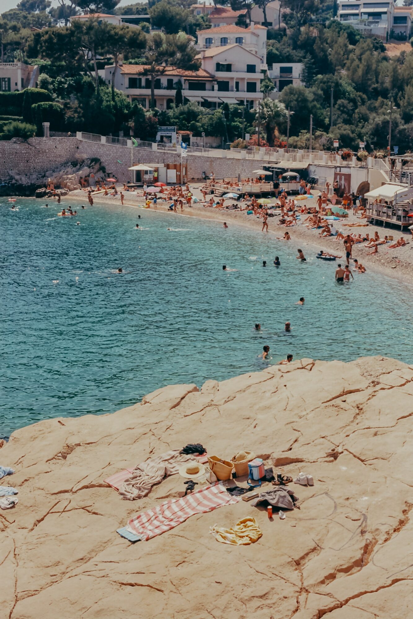 People sunbathe and swim on a rocky beach with buildings and trees in the background.