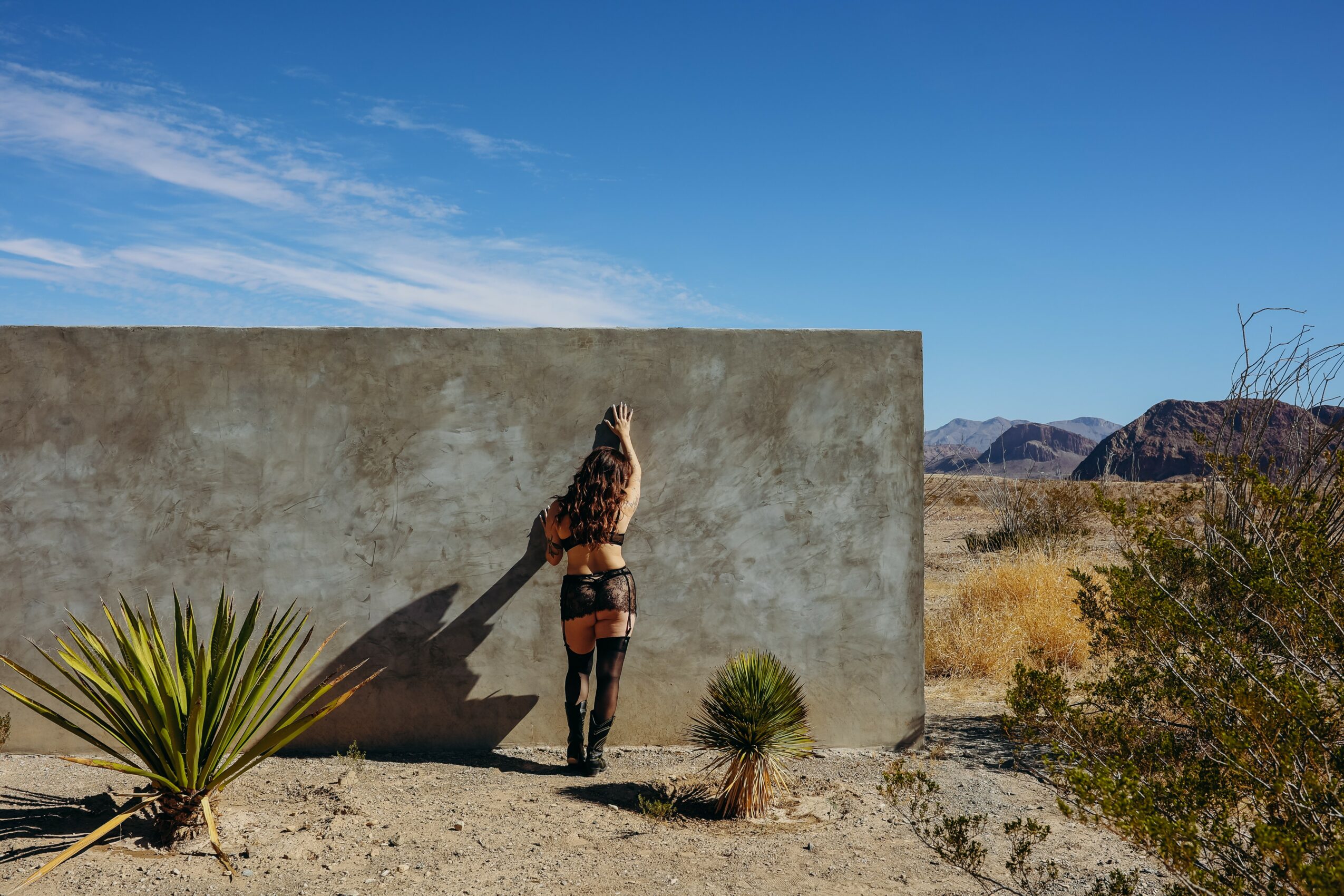 Person with long hair standing against a large concrete wall in a desert landscape, with mountains in the background and a clear blue sky.