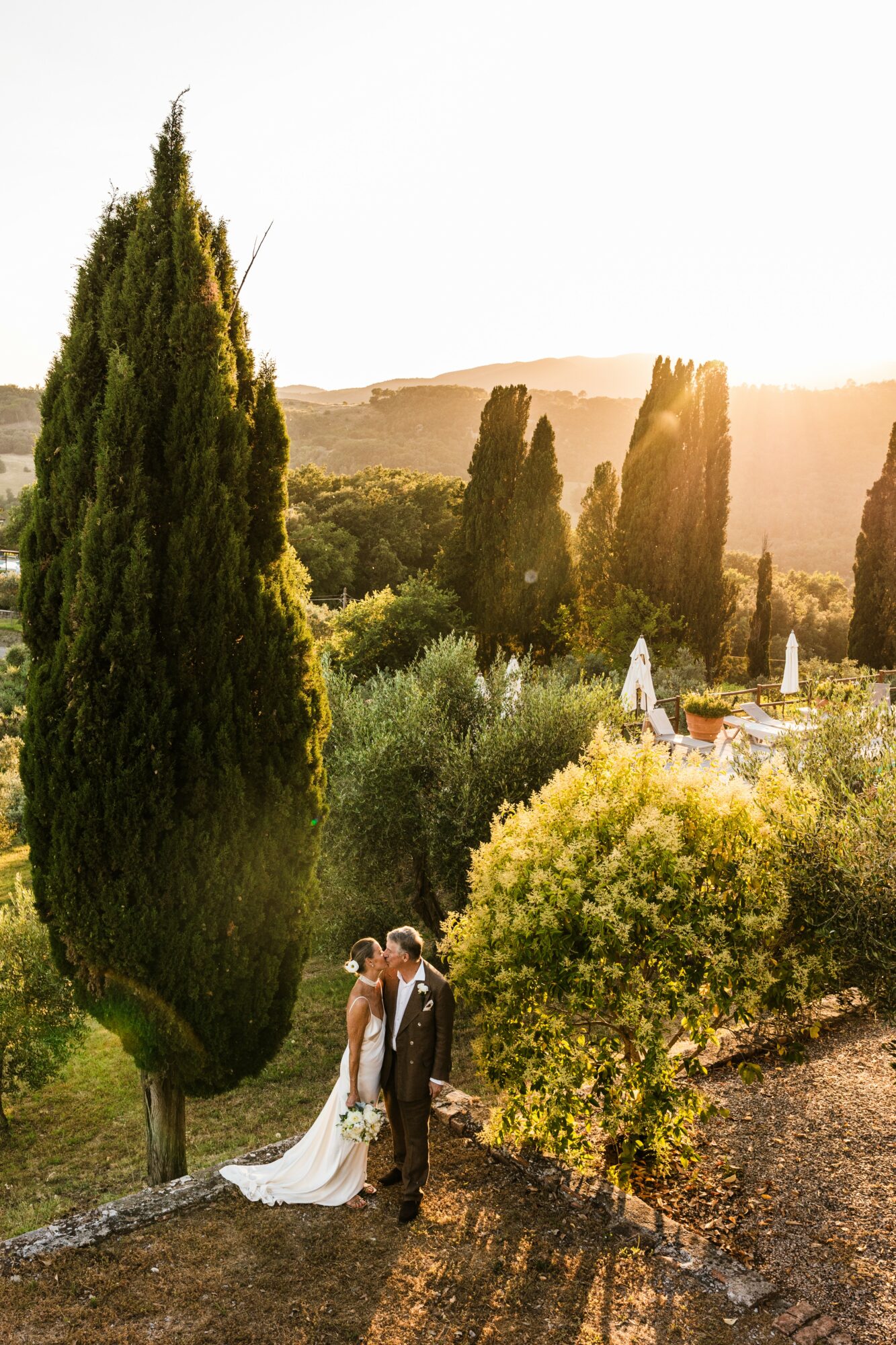 A couple in wedding attire standing outdoors among trees and greenery during sunset.