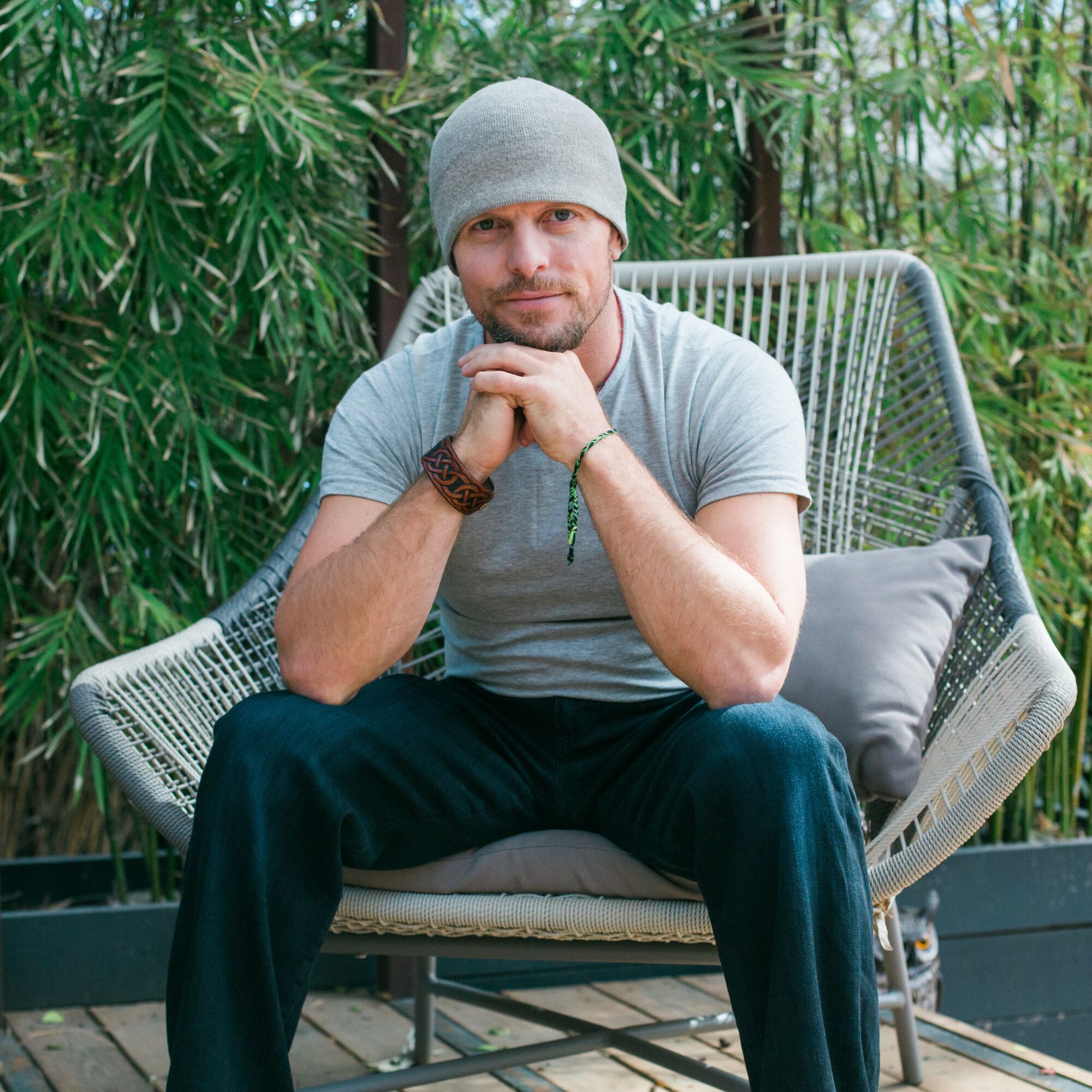 Man sitting on outdoor bench with greenery background, wearing a gray beanie and casual clothes, looking at camera.