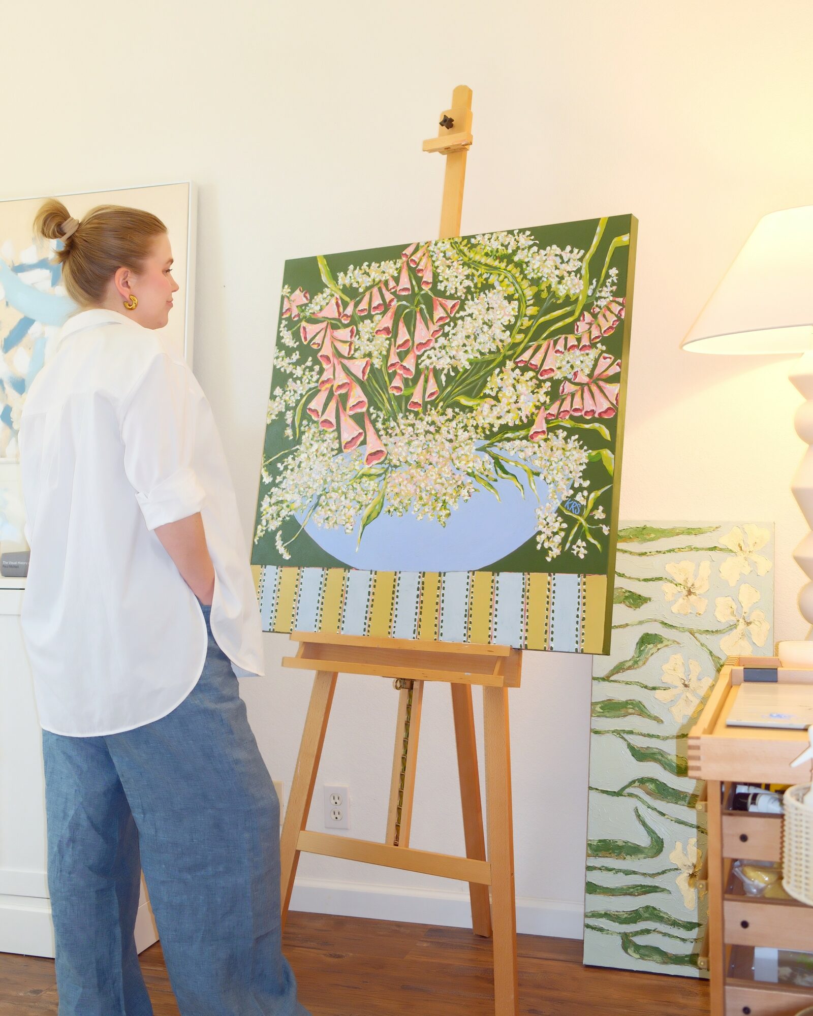 Person with hair in a bun wearing a white shirt and blue pants looking at a colorful painting on an easel in a room.